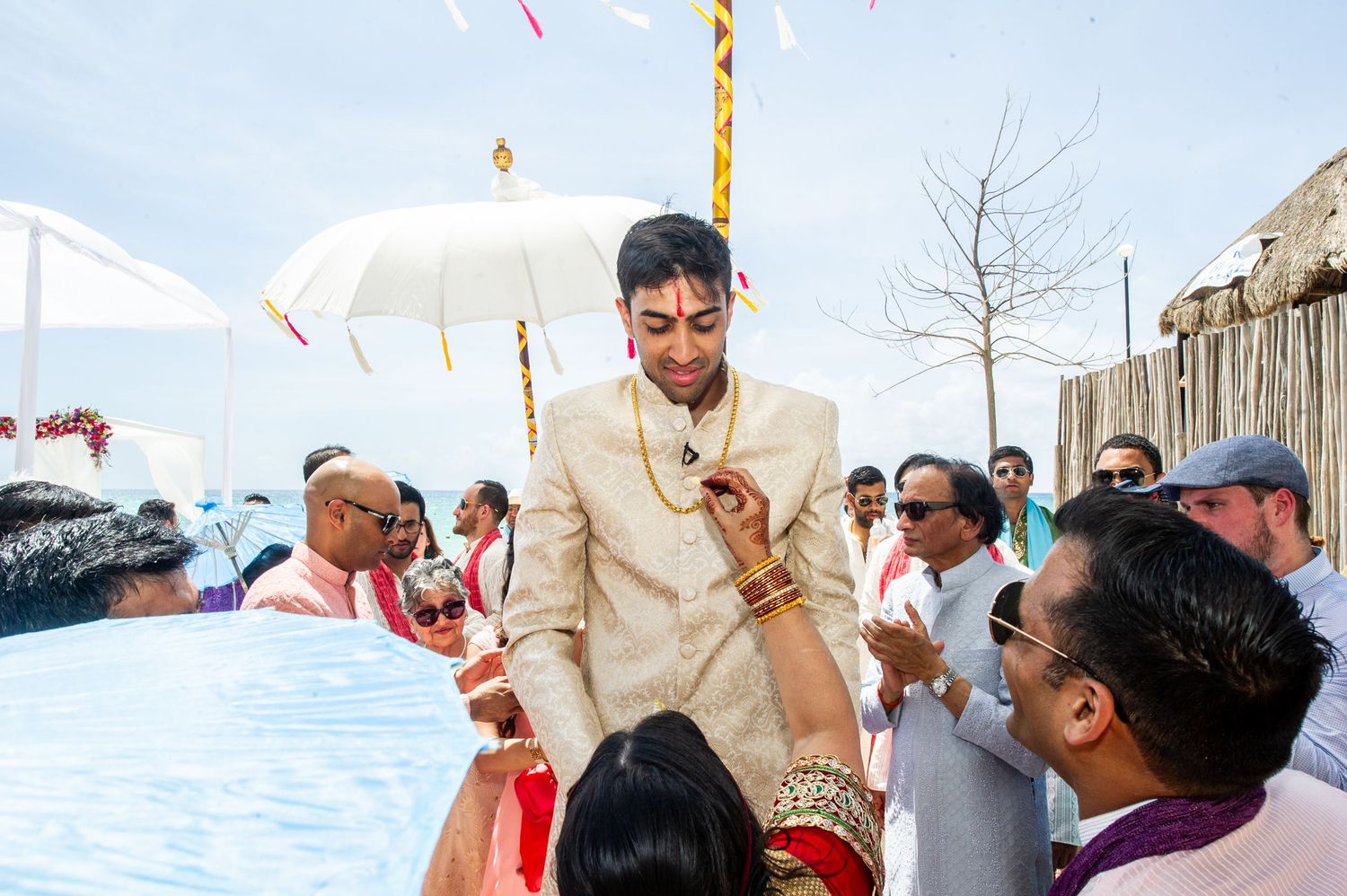 Groom in traditional Indian sherwani walks under white umbrella surrounded by wedding guests at outdoor ceremony.