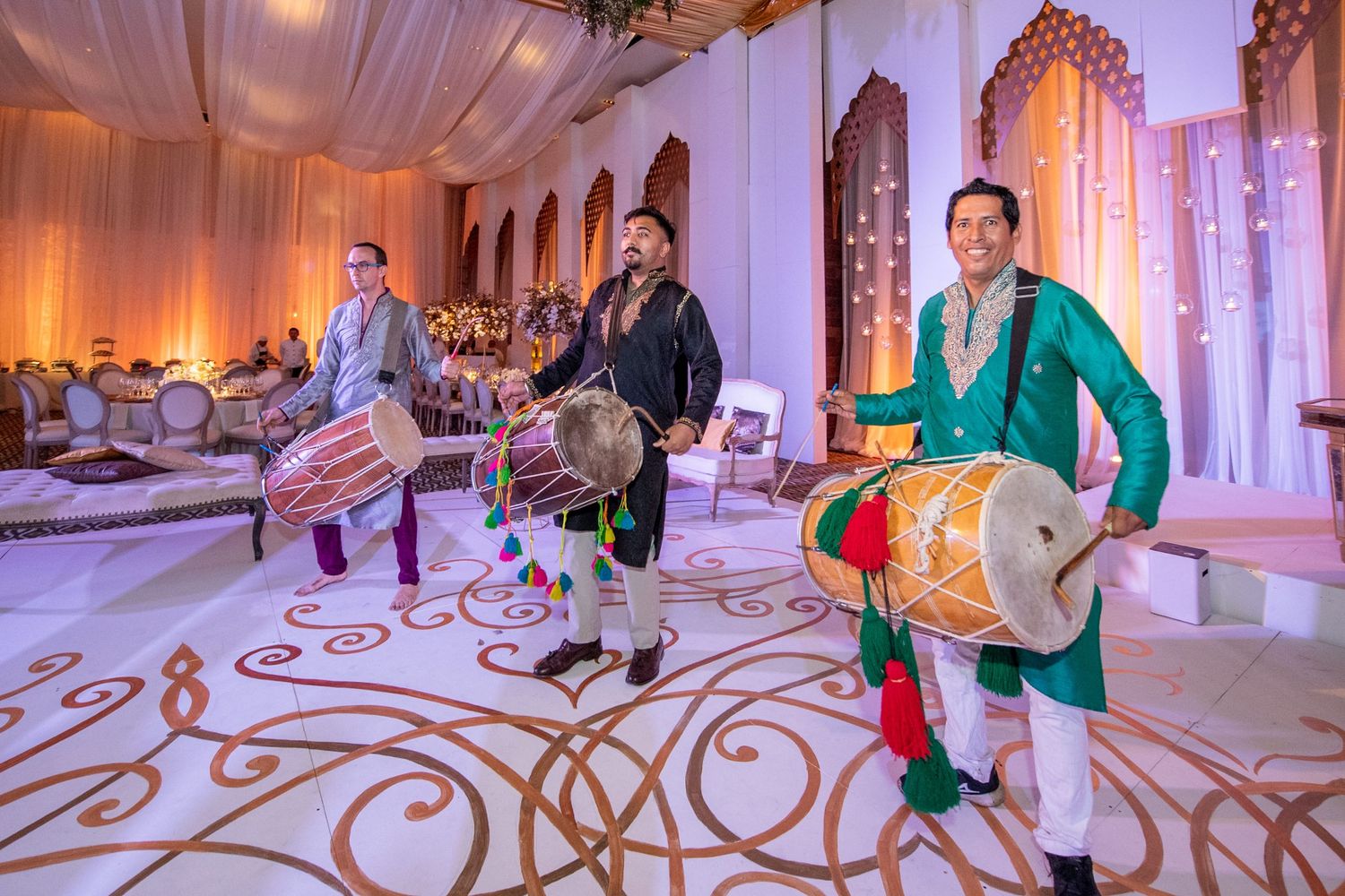 Three dhol drummers performing in vibrant traditional attire at an elegant Indian wedding celebration.