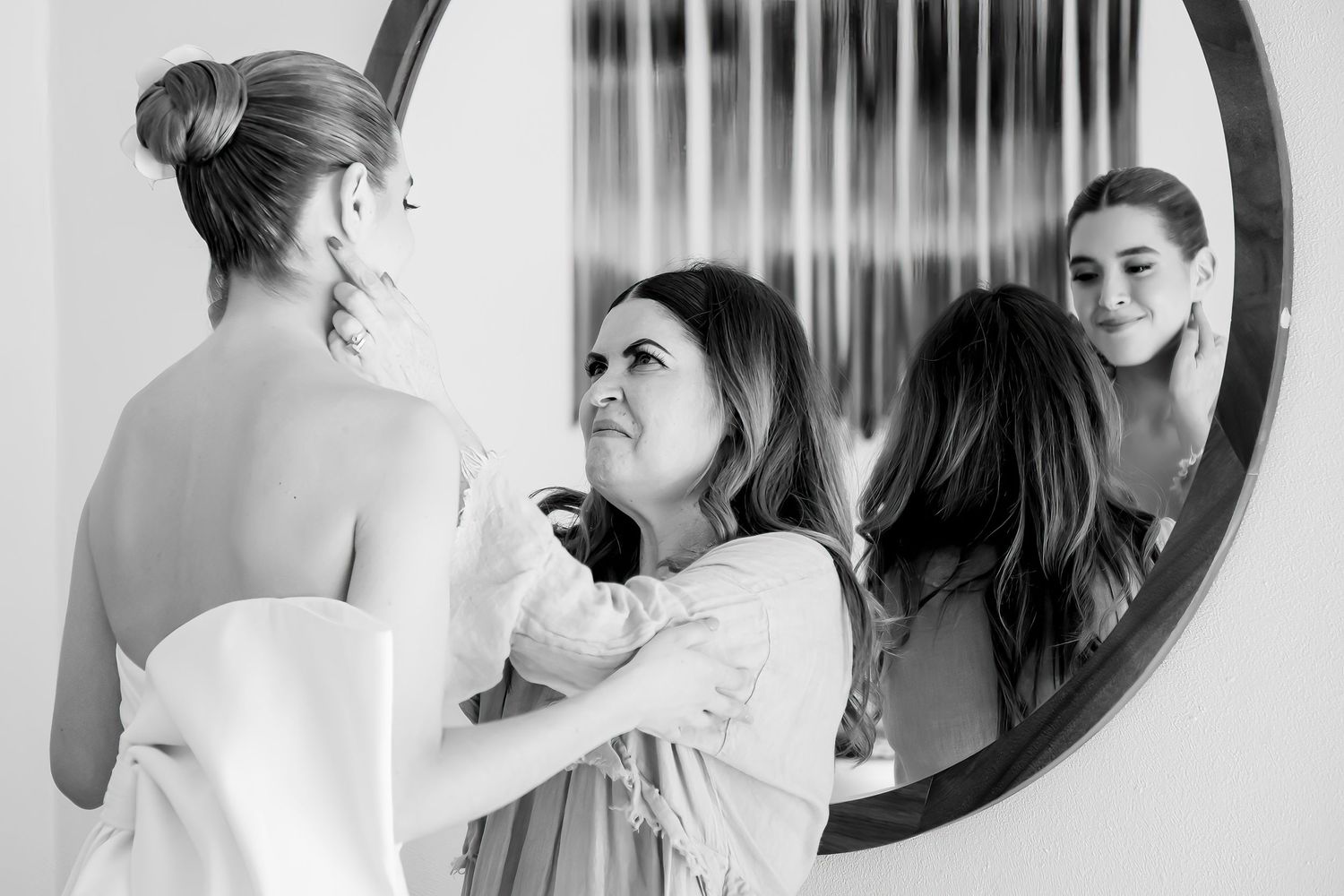 Black and white photo of bride being helped into wedding dress by two women near a round mirror.