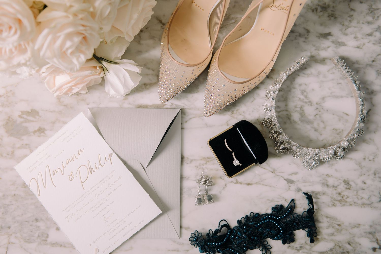 Elegant wedding flat lay with glittery heels, pearl earrings, crystal necklace, lace garter, and calligraphy invitation on marble.