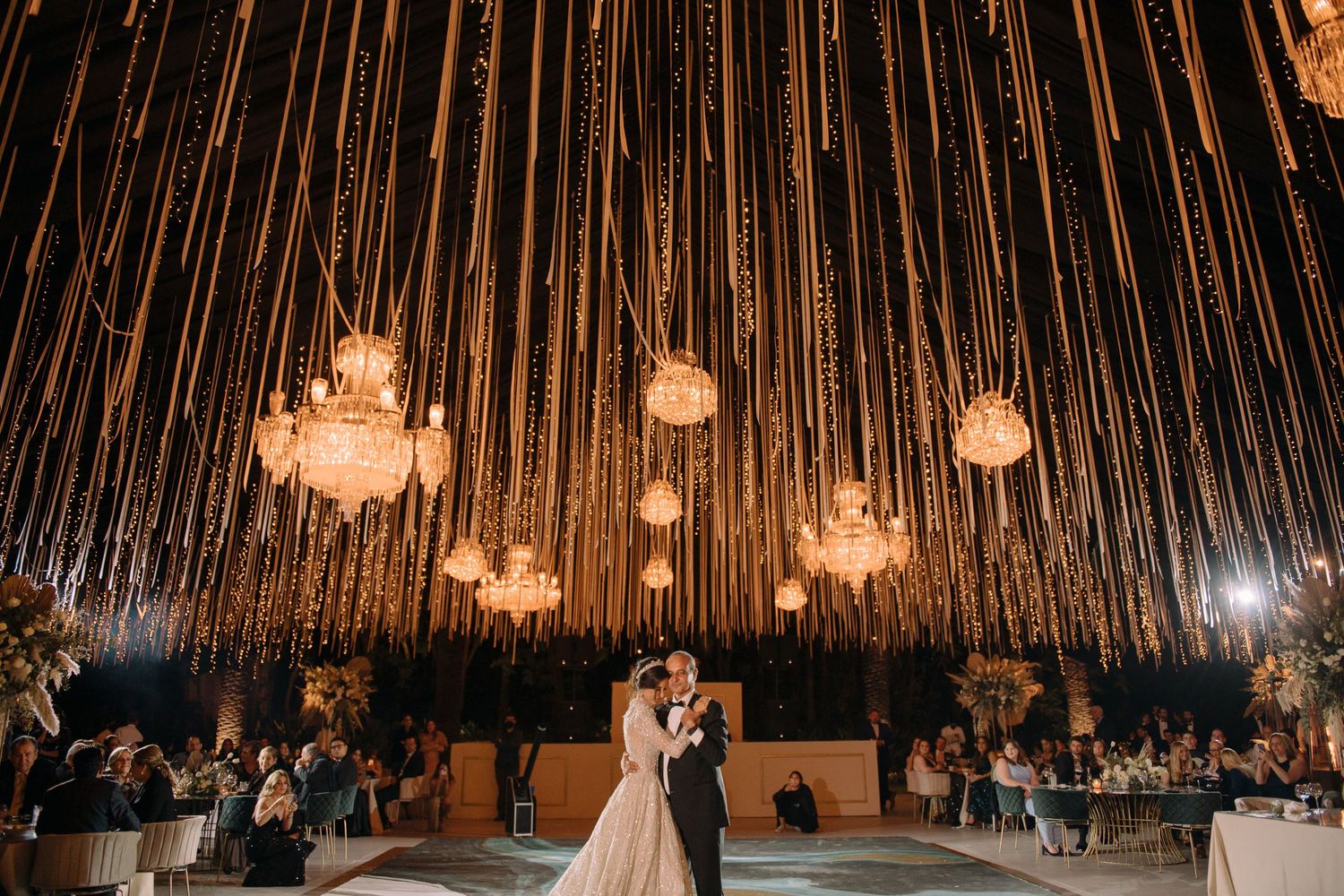 Bride and groom share first dance under glowing chandeliers and cascading string lights in elegant wedding reception venue.