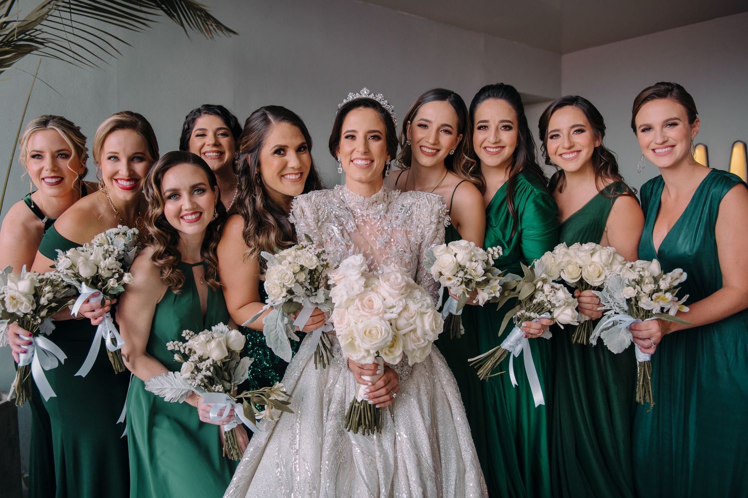 Bride in lace gown with tiara poses with bridesmaids in emerald green dresses holding white bouquets.