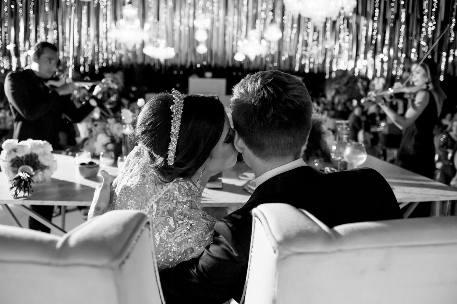Black and white photo of a bride and groom sharing a kiss at their wedding reception with elegant decor.