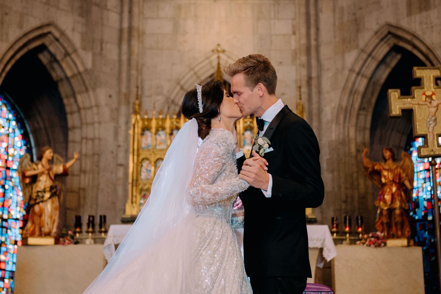 Bride and groom share first kiss at ornate church altar with stained glass windows on wedding day.