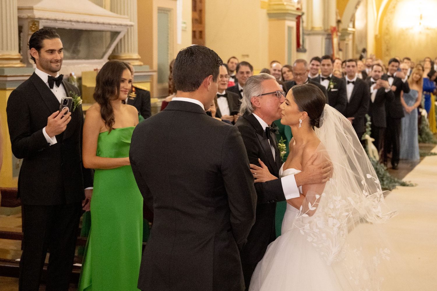 Bride in white lace gown and veil exchanges vows with groom in black tuxedo at elegant church wedding ceremony.