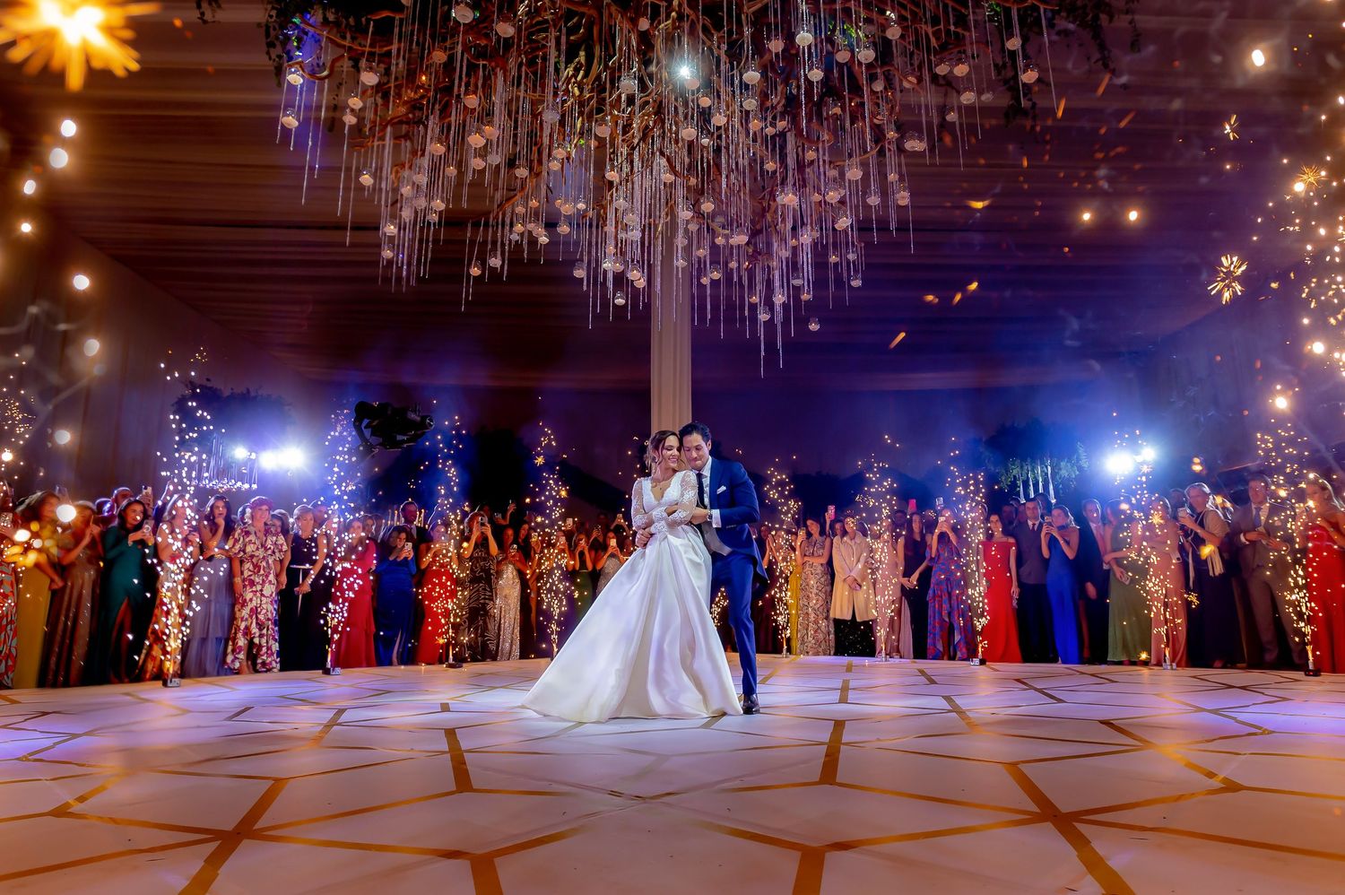 A grand wedding first dance under sparkling chandeliers with guests watching in the background.