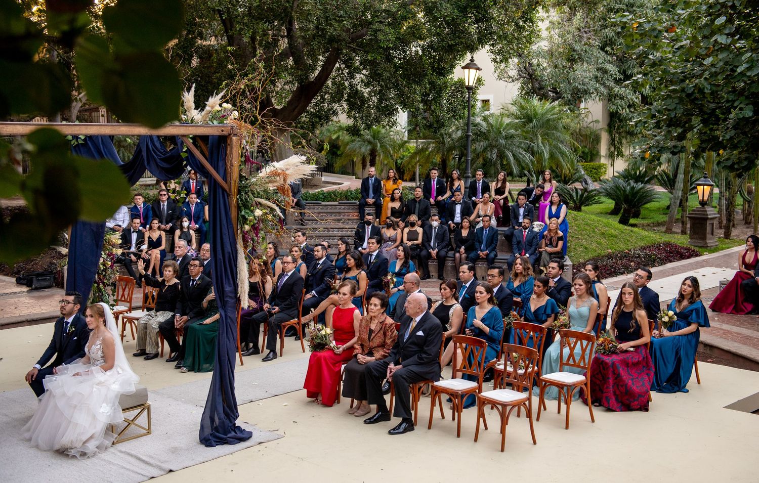 Elegant outdoor wedding ceremony with guests seated in colorful formal attire beneath a floral chuppah in a garden setting.
