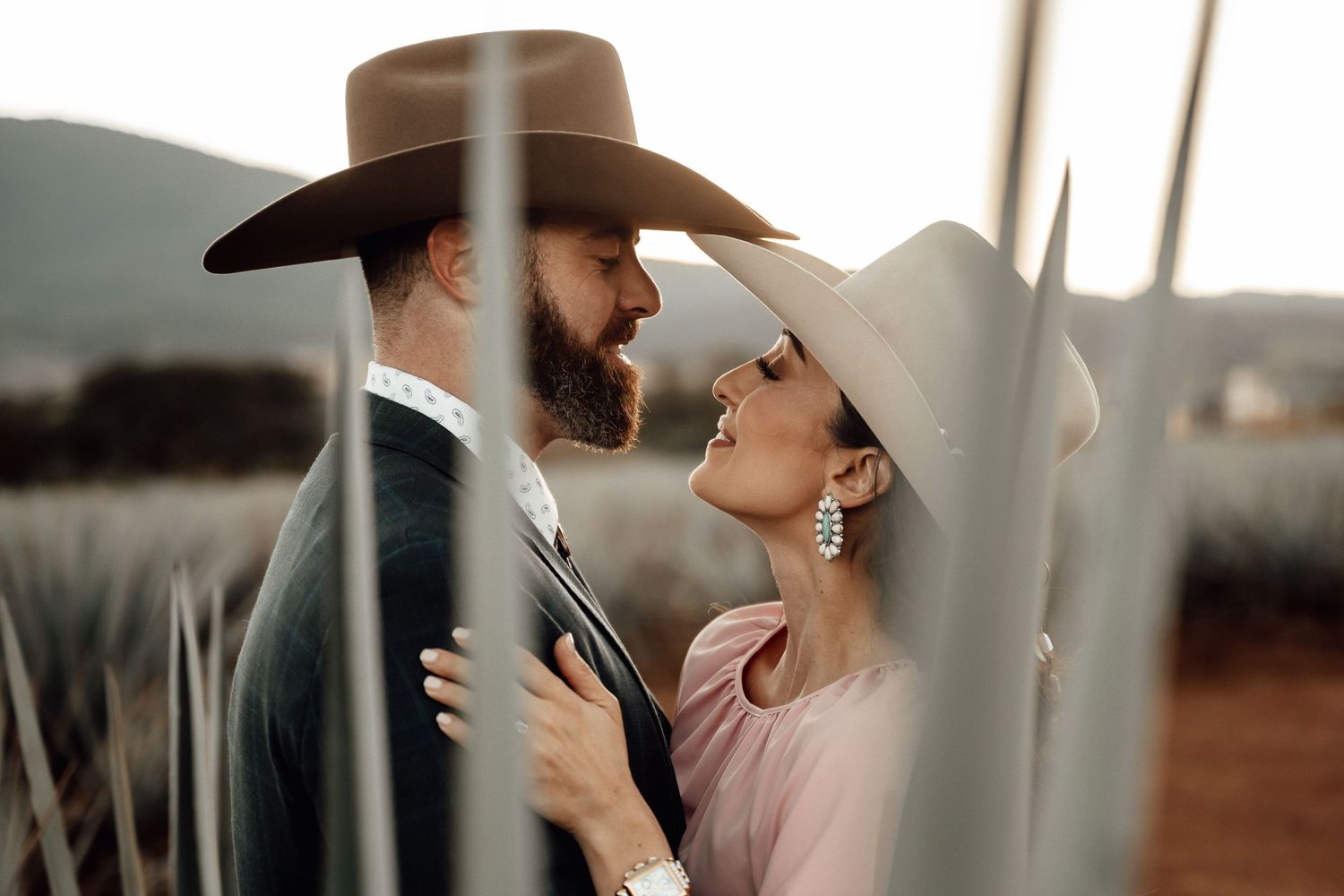 Couple in cowboy hats sharing a tender moment among agave plants at dusk.