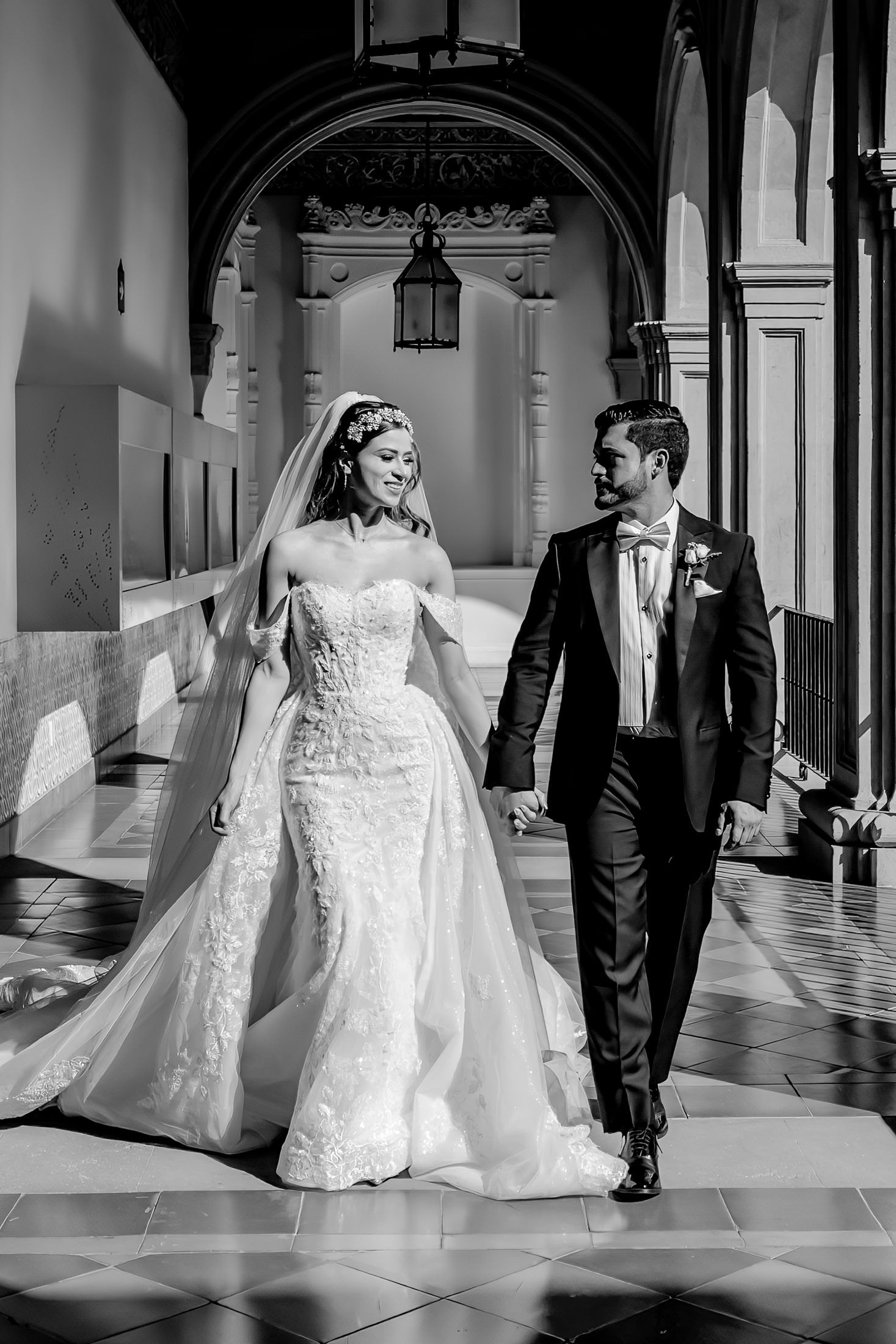 Black and white photo of bride and groom walking hand in hand through elegant arched hallway on wedding day.