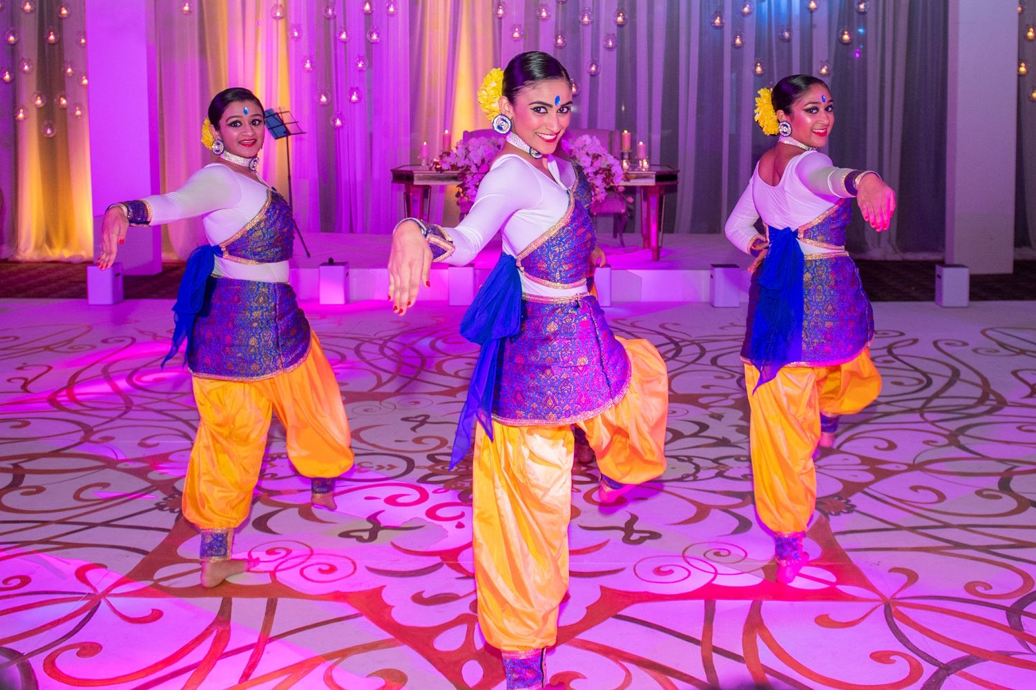 Three women performing a vibrant Bollywood dance in colorful blue and gold costumes on a pink-lit stage.