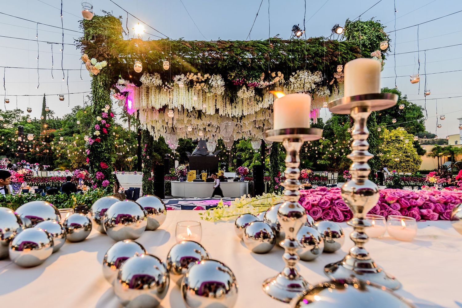 Elegant outdoor wedding reception with silver candelabras, mirrored orbs, pink florals, and a floral arch backdrop at sunset.