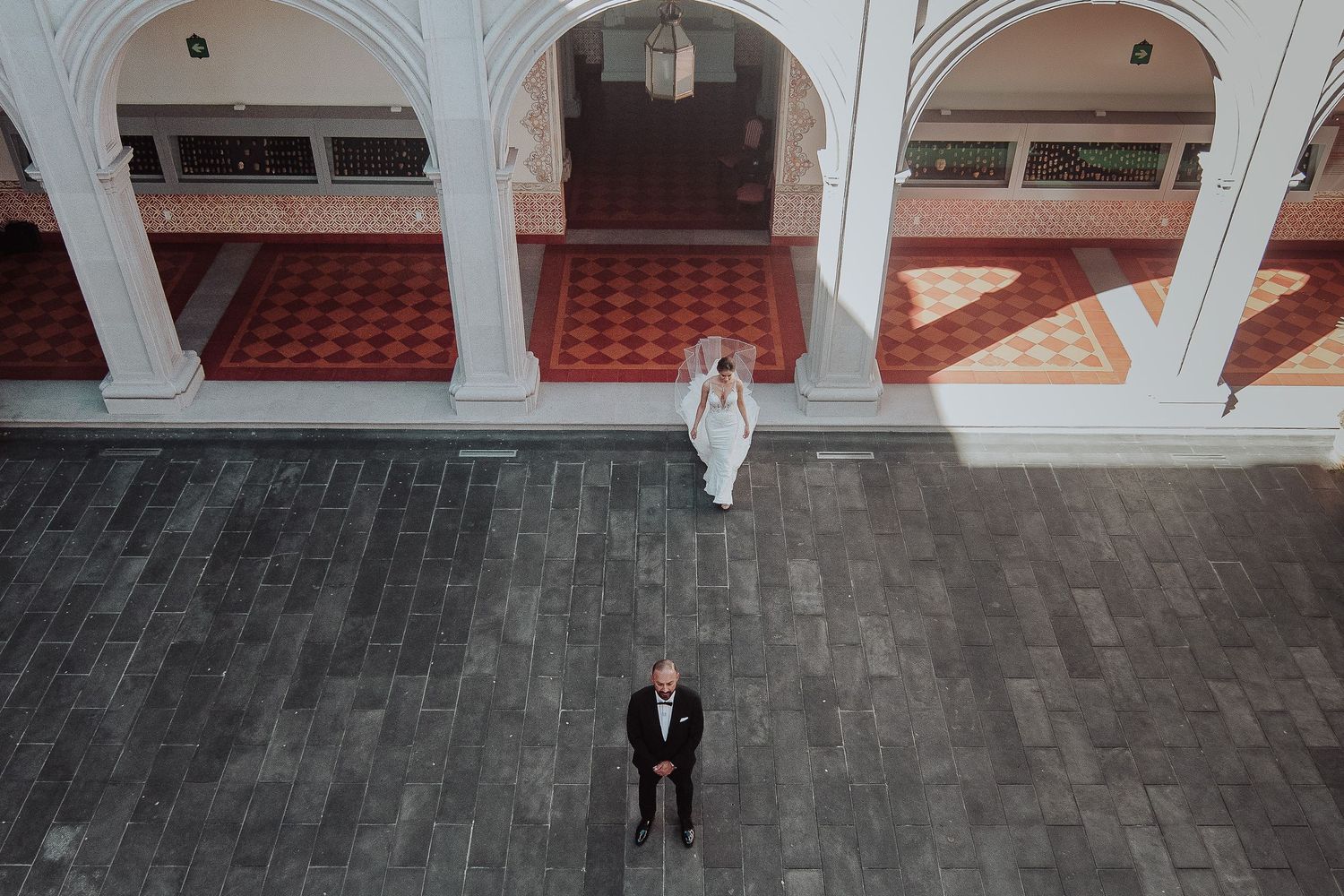 Aerial view of bride and groom standing apart in elegant arched venue with ornate tiled floors.