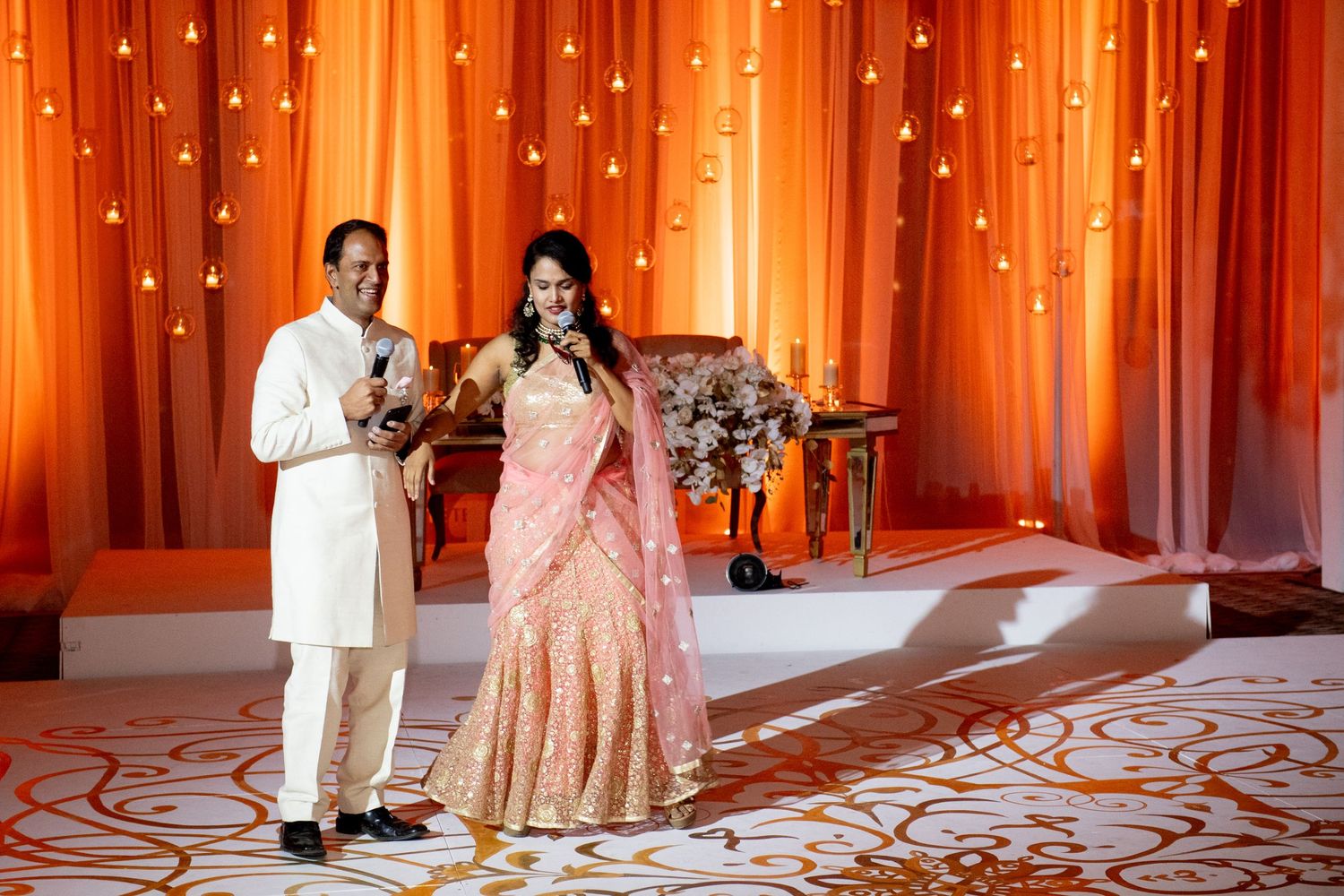 Couple in traditional Indian attire speaking at a wedding reception with orange floral backdrop.