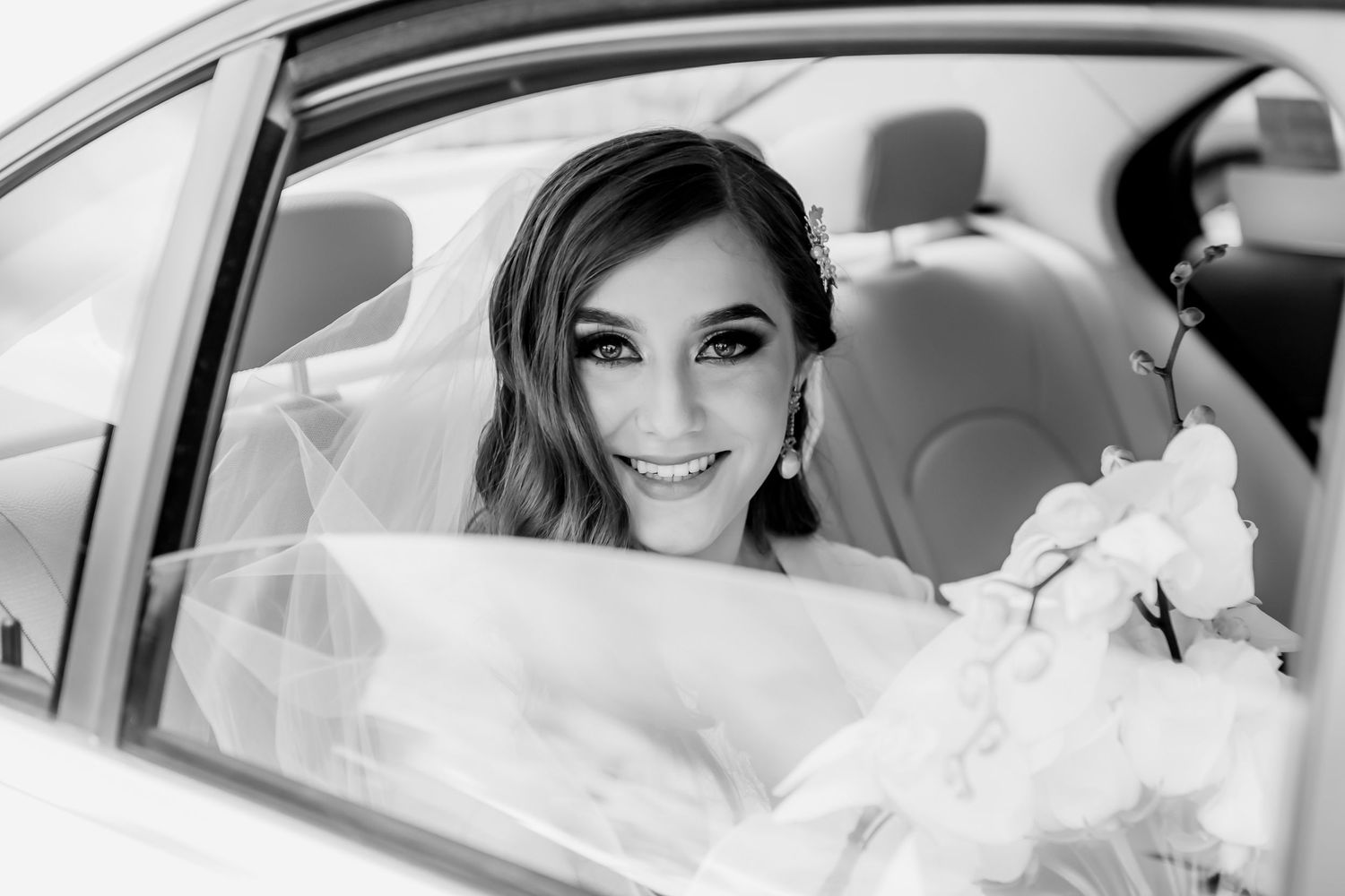 A black and white portrait of a bride smiling from inside a wedding car.