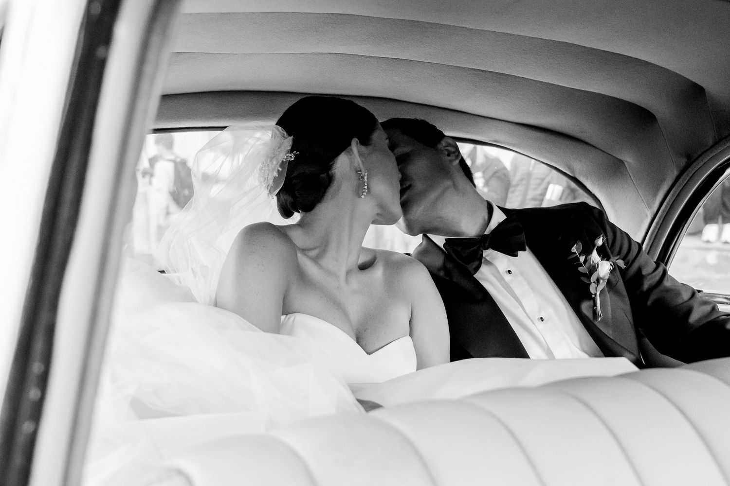 Black and white photo of newlyweds sharing a tender kiss in the backseat of a vintage car.