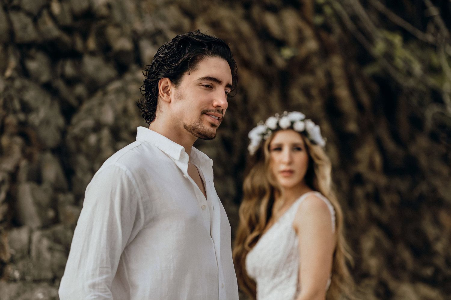 A groom in a white shirt stands before rocky cliffs with a bride wearing a floral crown softly blurred behind him.