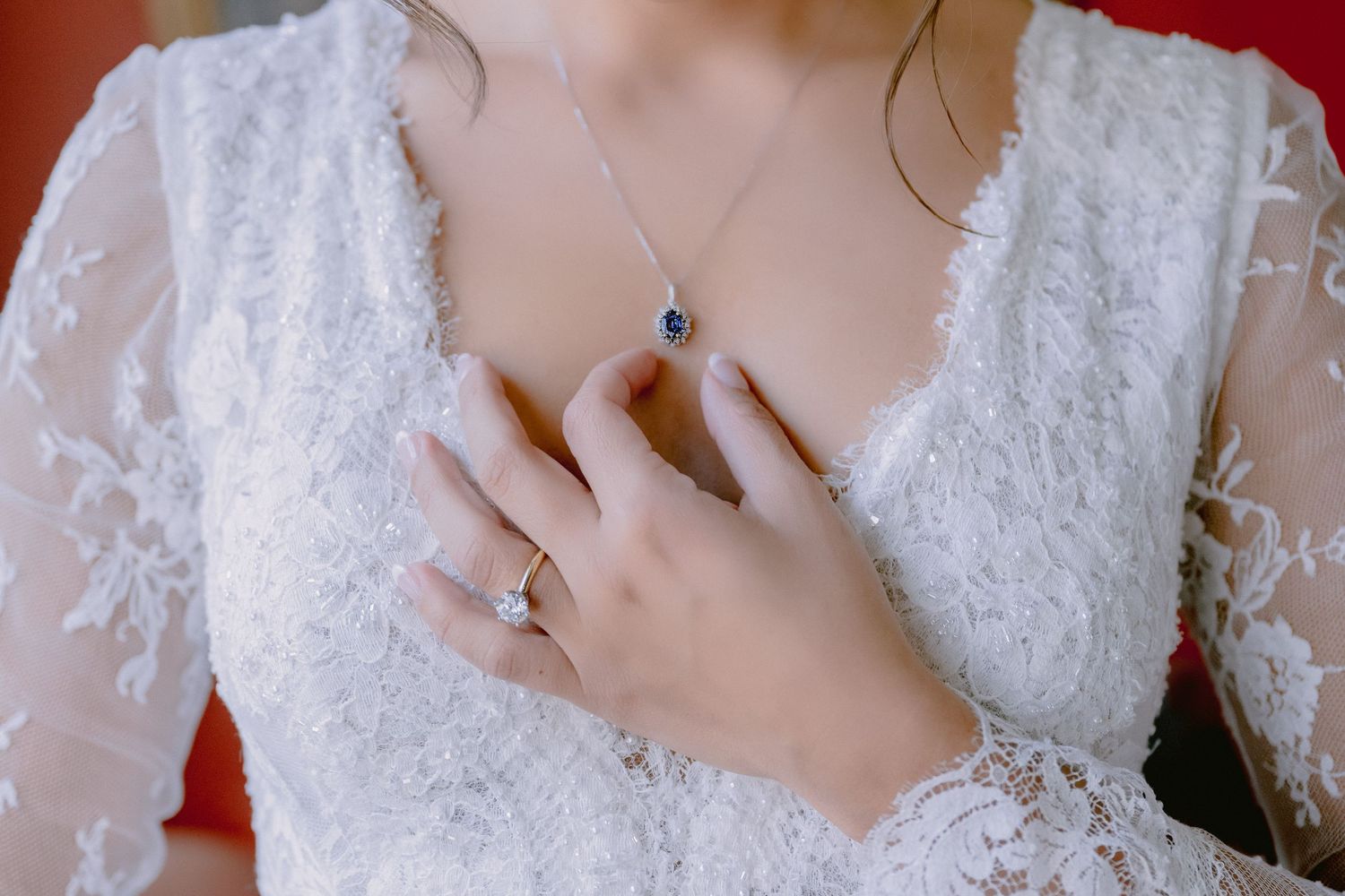 Close-up of delicate white lace wedding dress bodice with sapphire necklace and engagement ring.