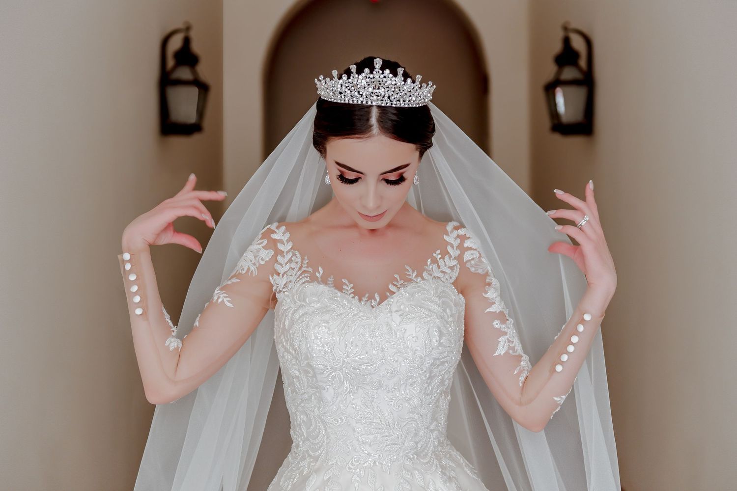 A bride adjusts her veil while wearing a tiara and detailed lace wedding dress.