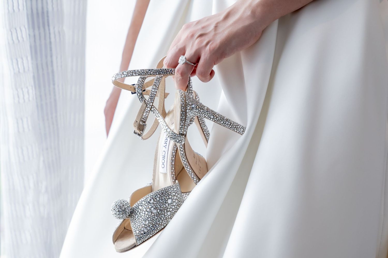 Hands holding sparkly silver wedding shoes against white fabric background.