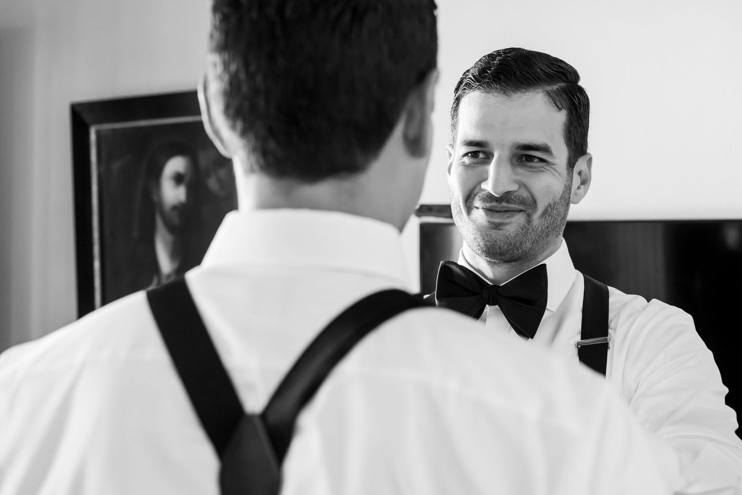 Black and white photo of two grooms in suspenders and bow ties sharing a smile during wedding preparations.