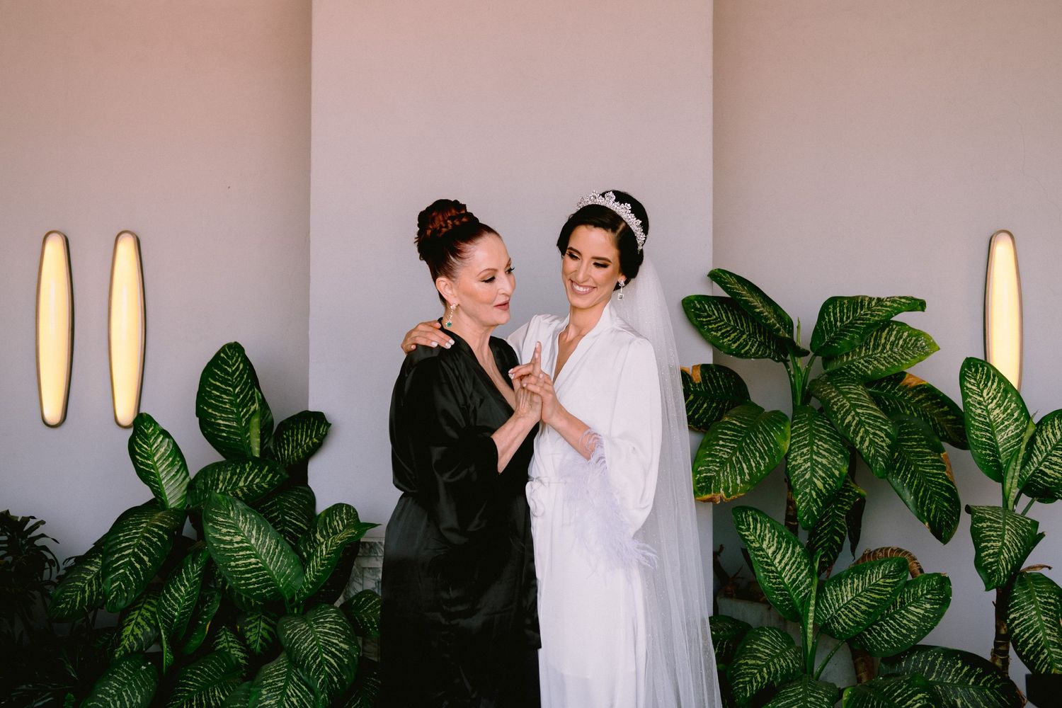 Bride in white gown with bridesmaid in black robe sharing a moment surrounded by tropical plants.