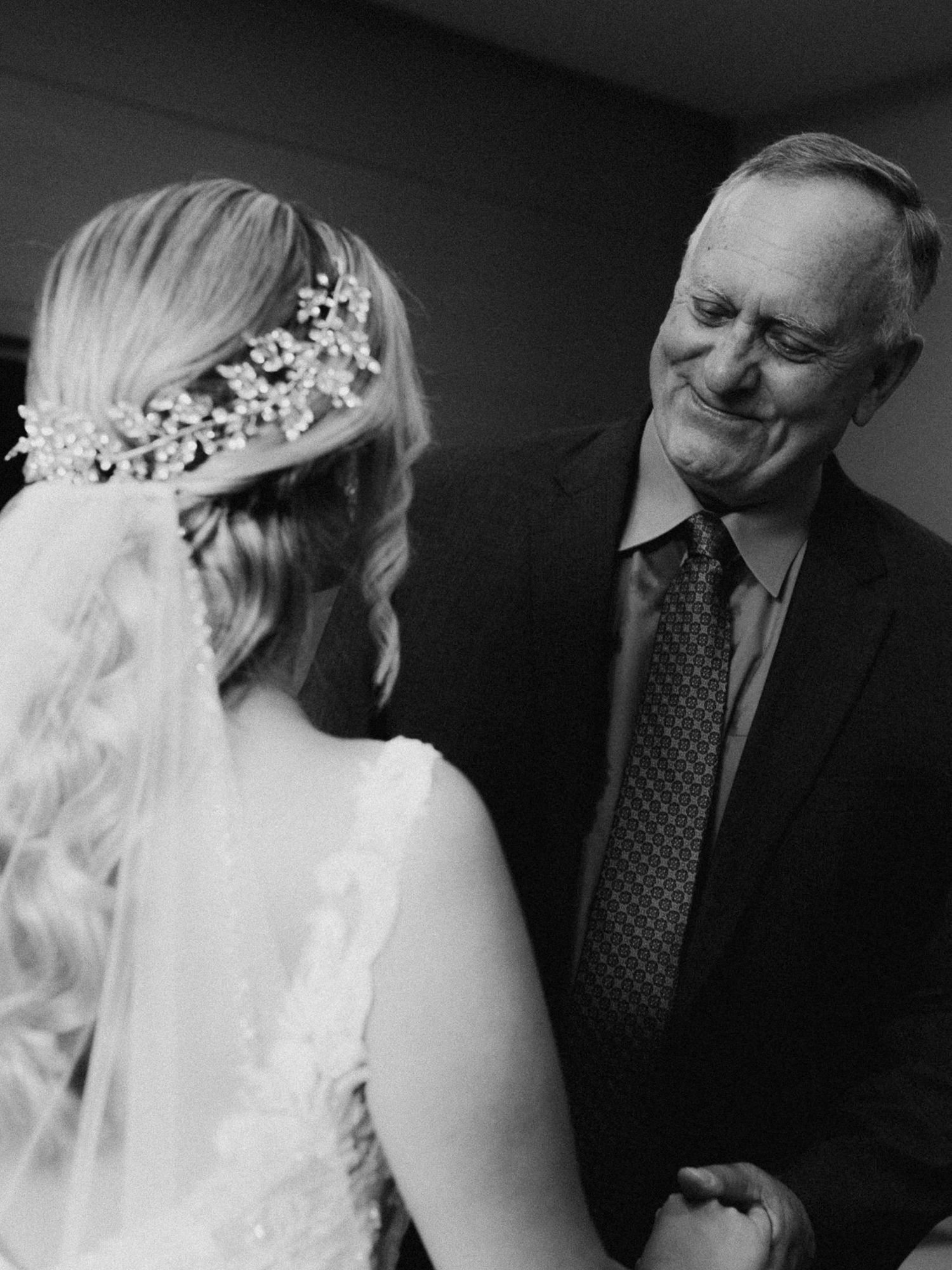 Black and white photo of a bride in a veil and hair piece sharing an emotional first look moment with her father.