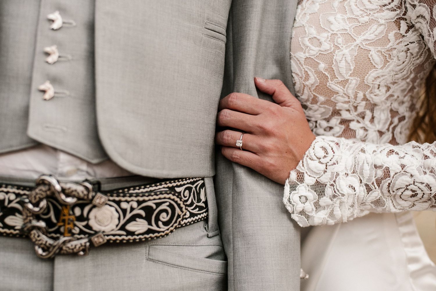 Close-up of bride and groom in lace wedding dress and gray suit with ornate belt, bride wearing diamond ring.
