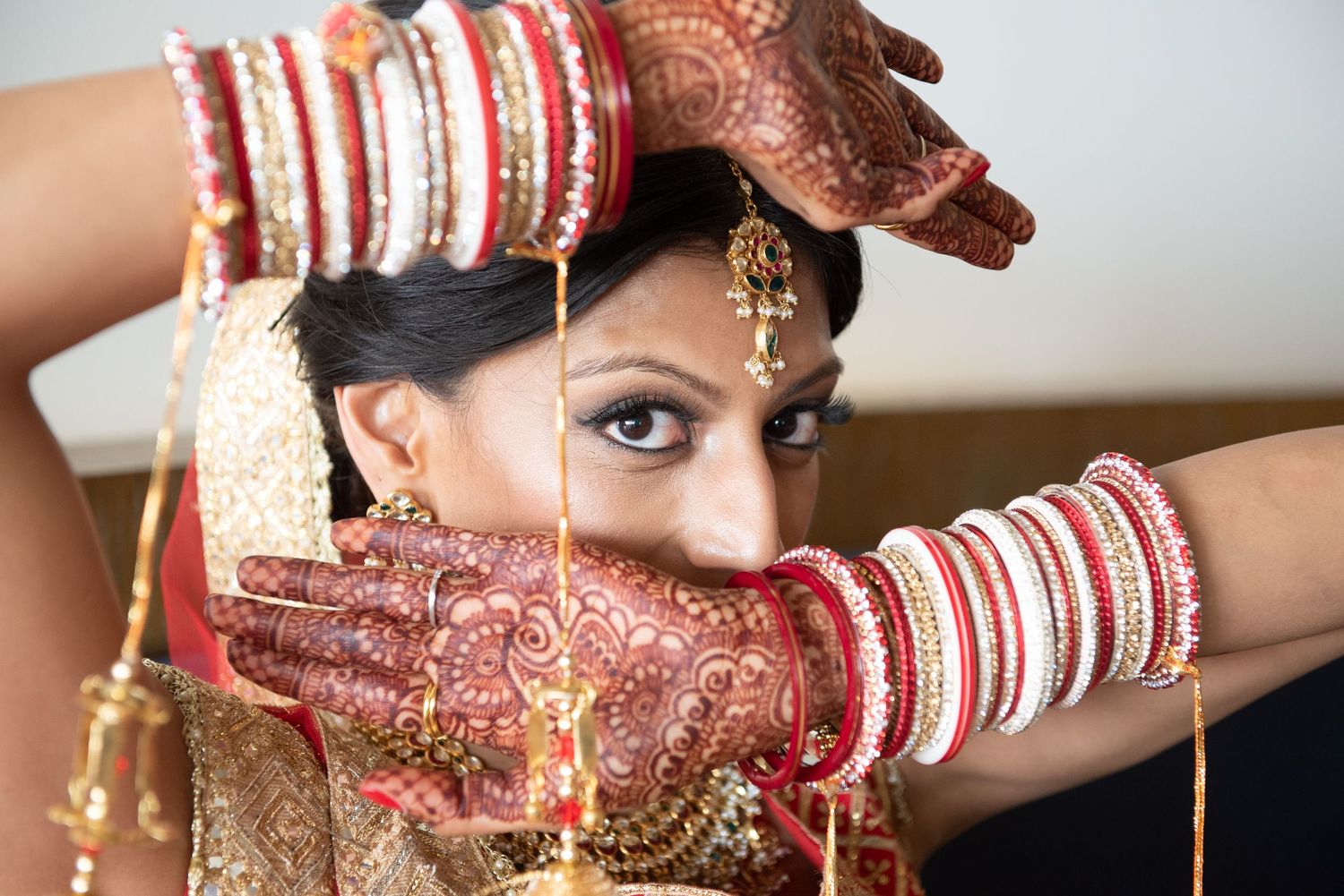 Indian bride in traditional red bridal attire with mehndi henna hands, gold jewelry, and bangles posing elegantly.