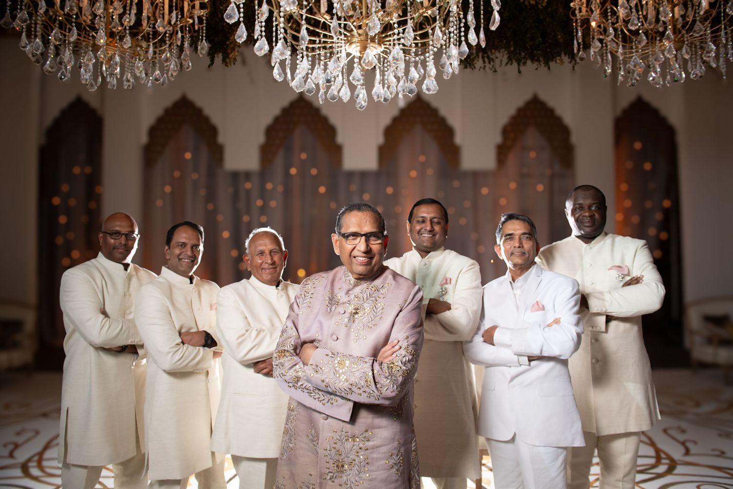 Group of men in elegant white traditional Indian attire posing under a crystal chandelier in an ornate venue.