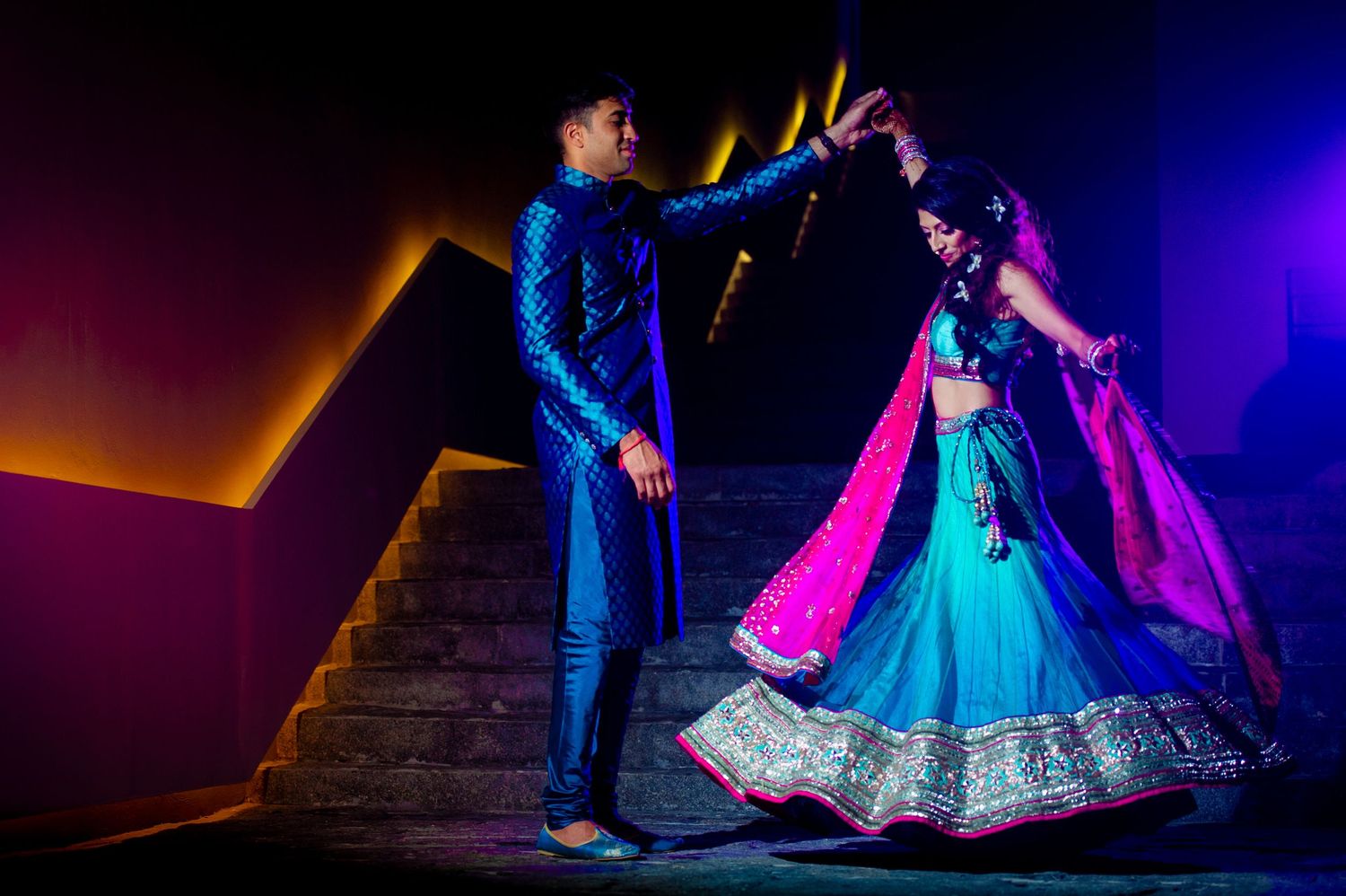Couple in vibrant blue and pink traditional Indian attire dancing elegantly on stone steps under colorful lighting.