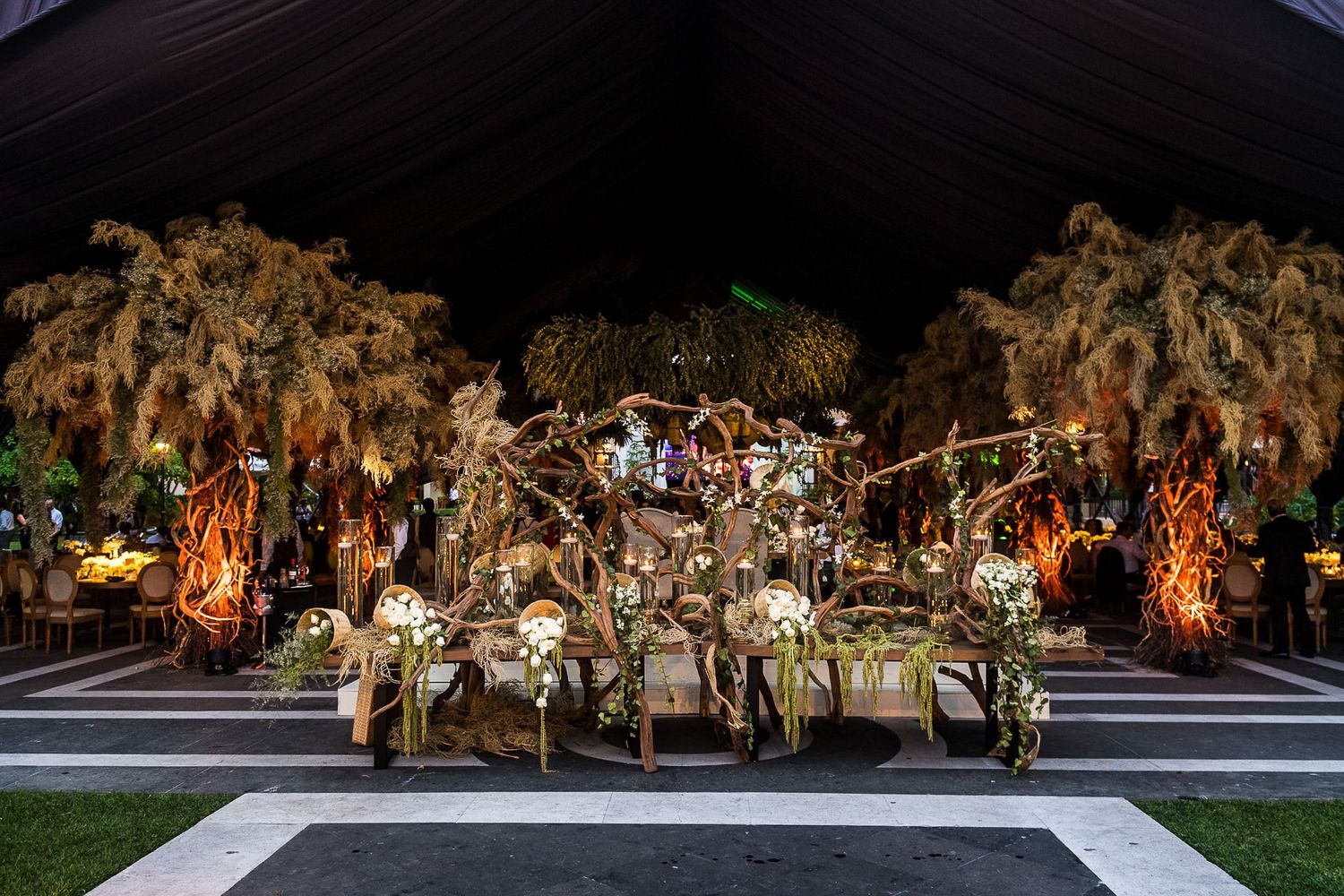 Elegant outdoor event setup with illuminated floral arrangements, draped trees, and ornate decor under a dark canopy at night.