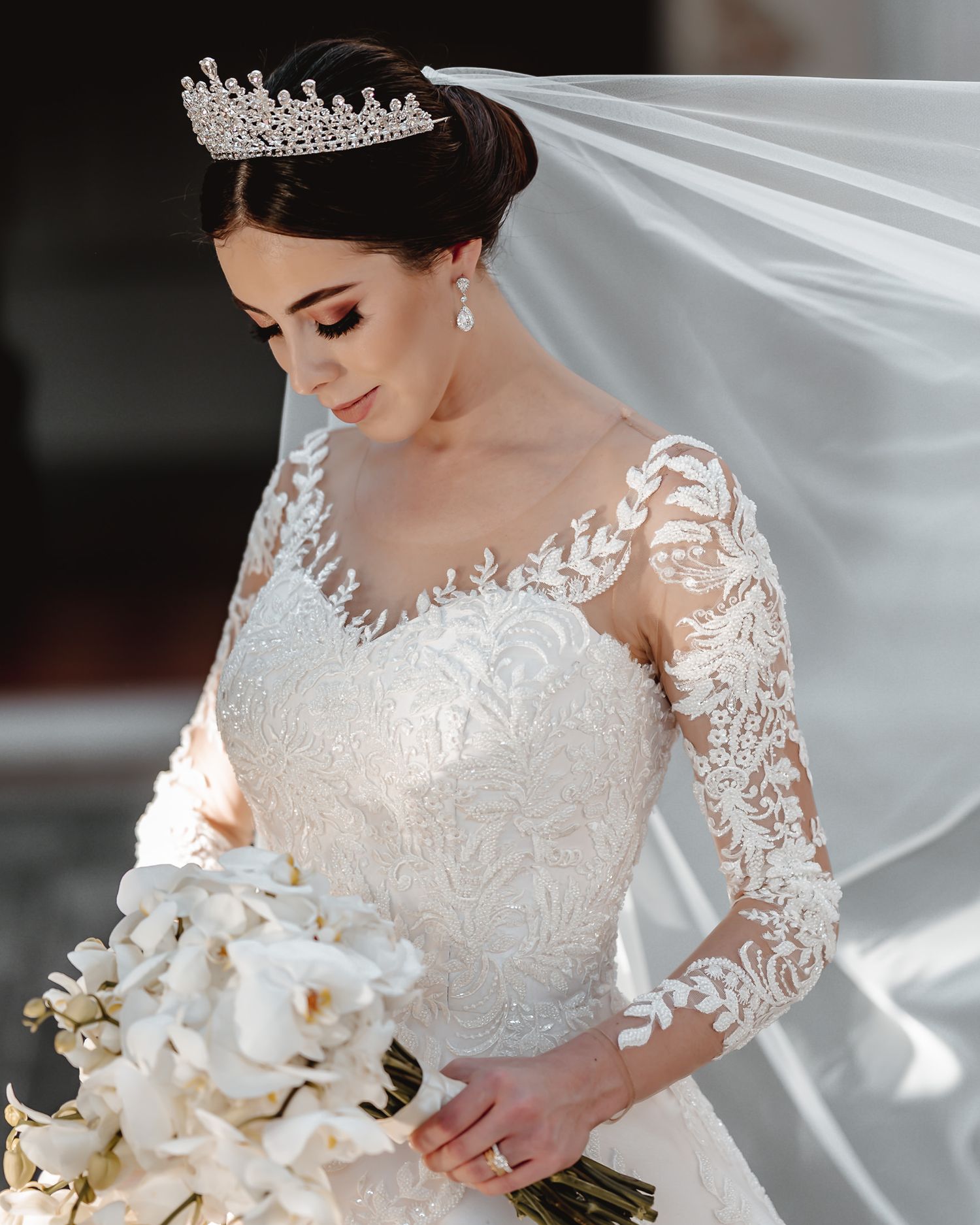 Elegant bride in lace long-sleeve wedding gown, crystal tiara, and veil, holding white orchid bouquet.