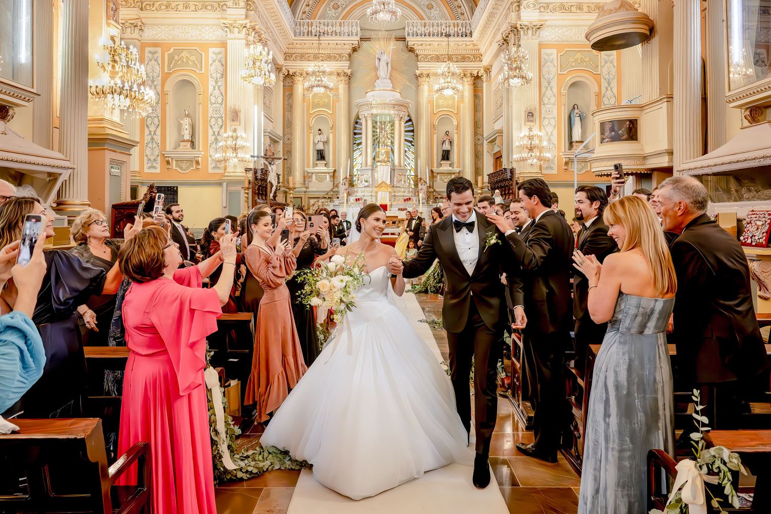 Bride and groom walk down church aisle surrounded by cheering guests during elegant wedding ceremony.