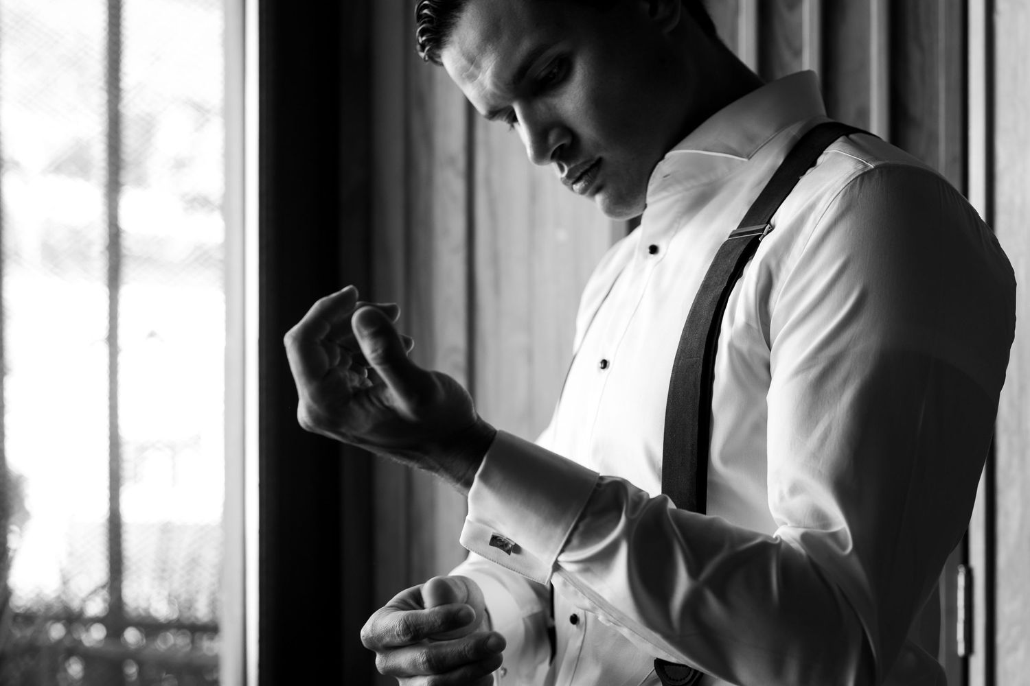 Black and white photo of a well-dressed man in white shirt and suspenders fastening his cufflinks by a window.