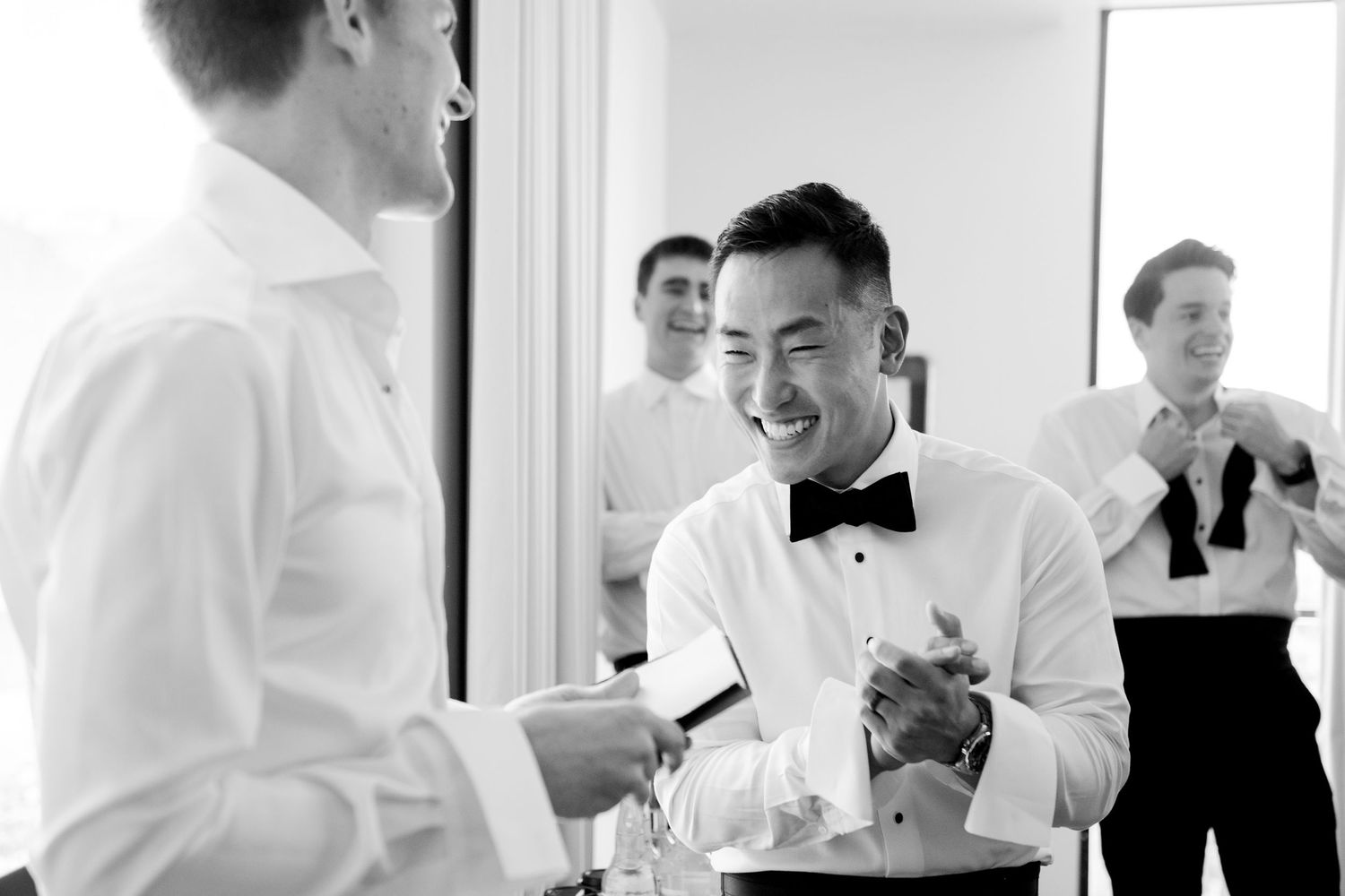 Groom in bow tie laughing with groomsmen during wedding preparations in black and white photo.