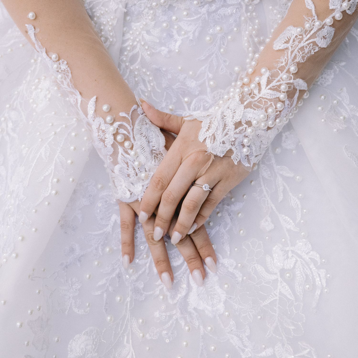 Close-up of hands with engagement ring resting on intricate beaded white wedding gown.