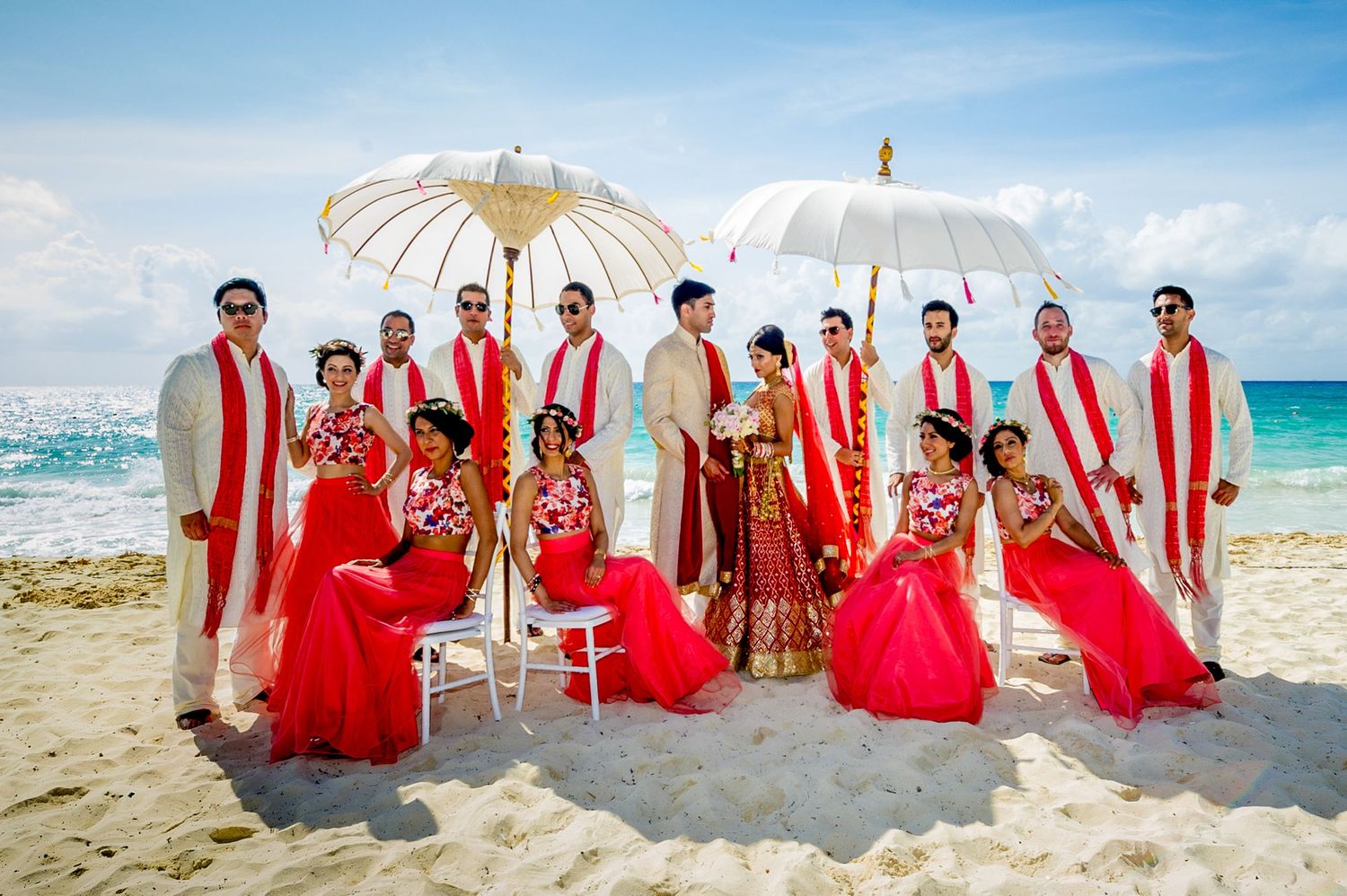 Indian wedding party in red and gold attire posing on a tropical beach with white parasols and ocean backdrop.