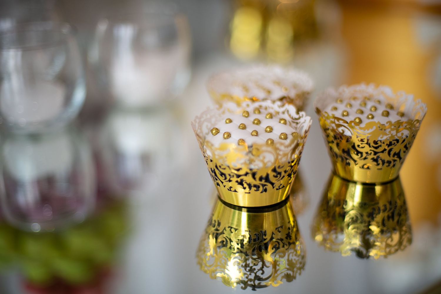 Elegant gold filigree decorative cups on a reflective surface with blurred glassware in the background.