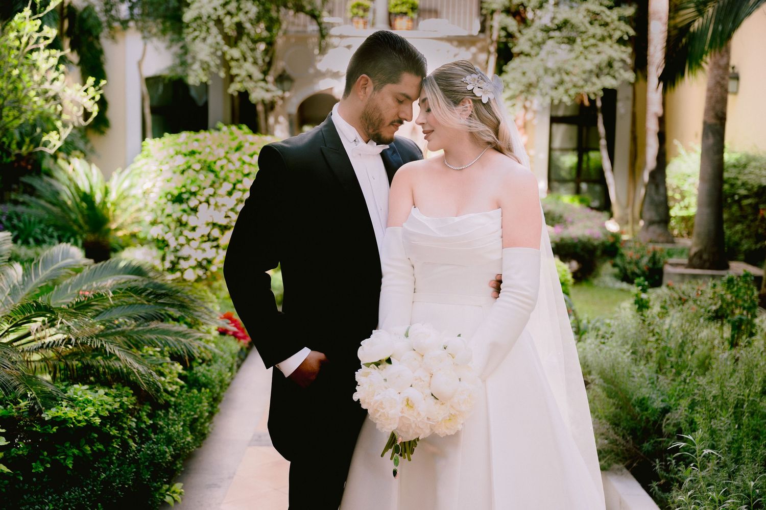 Bride and groom share an intimate moment in a lush garden, she holds white bouquet wearing strapless gown with gloves.