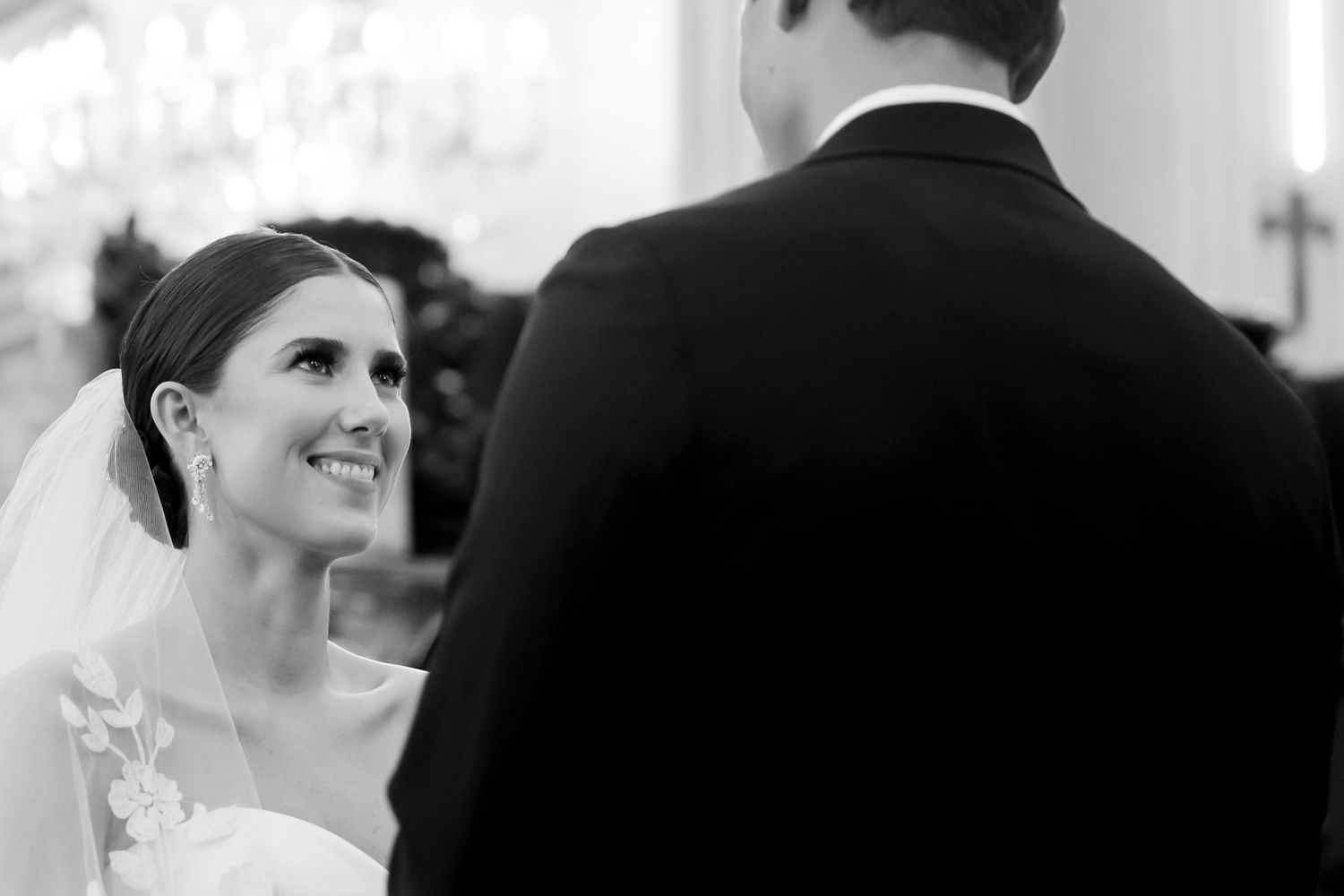 Black and white wedding photo of a smiling bride in veil looking up at groom during ceremony.