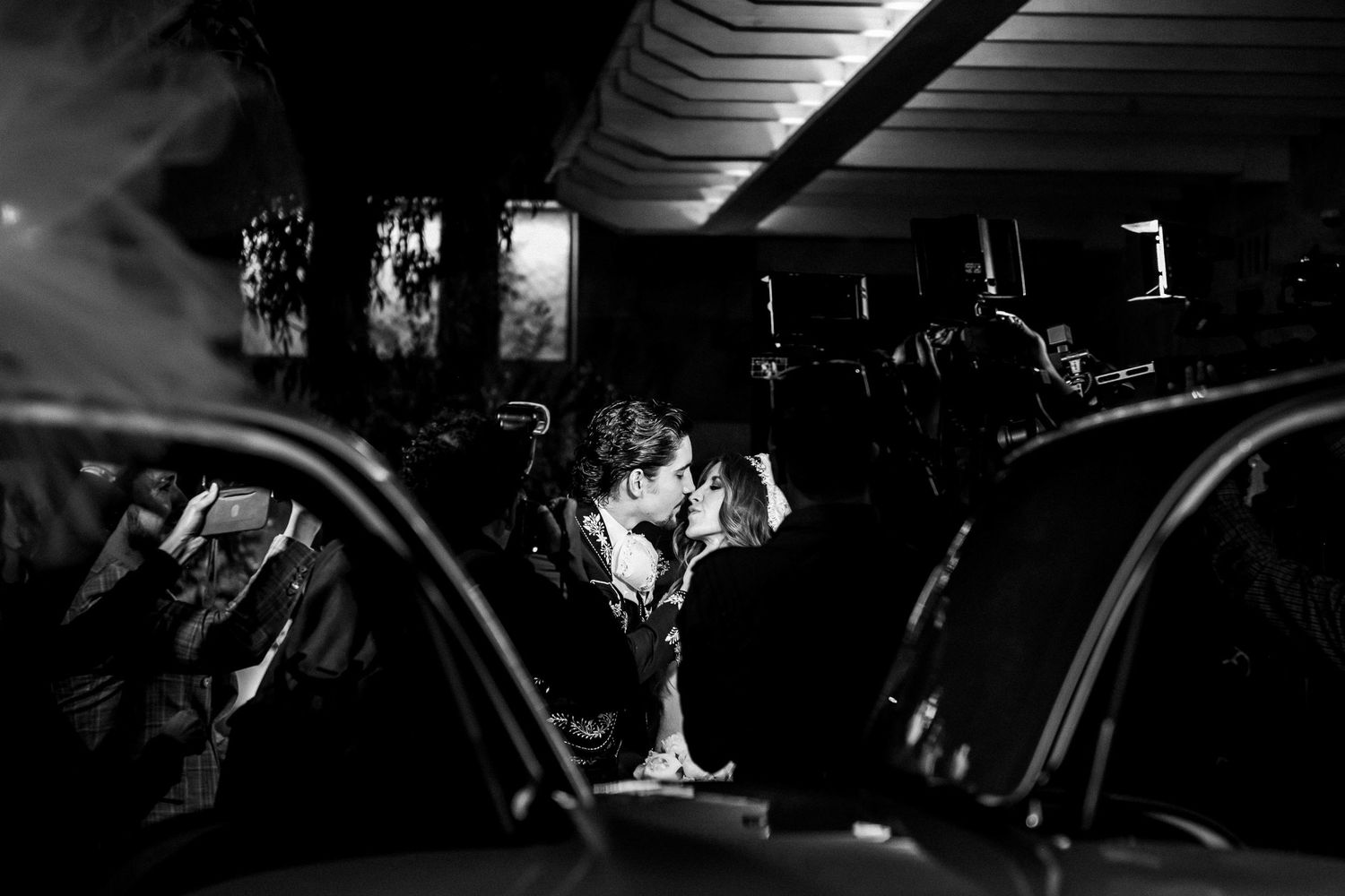 Black and white photo of a couple sharing a romantic kiss between vintage cars at night.