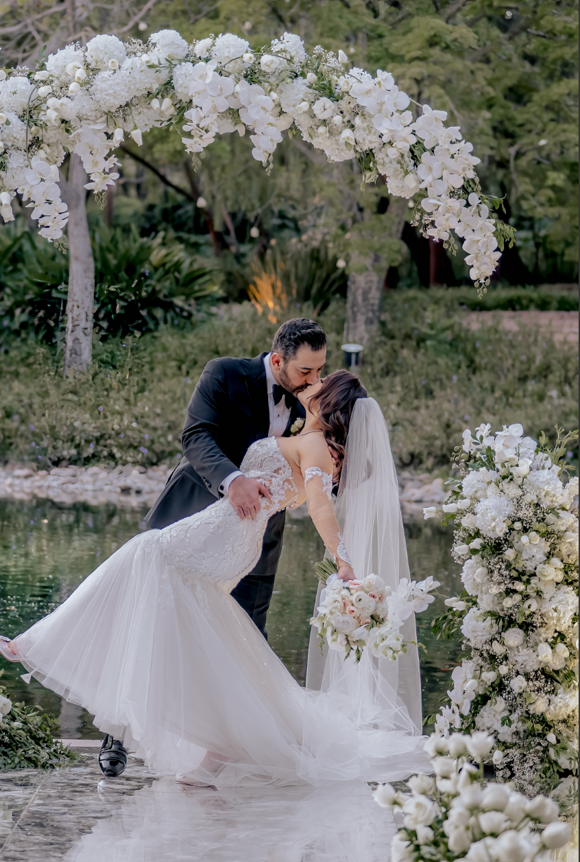 Romantic wedding couple sharing a kiss under an arch of white orchids by a pond.