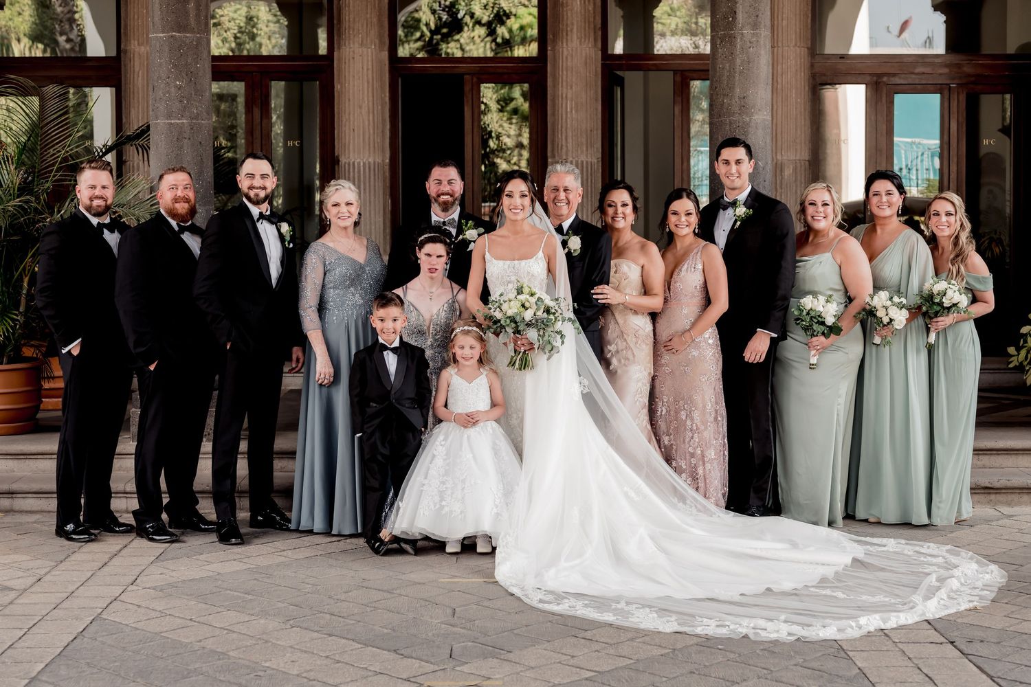 Wedding party poses together in formal attire with bridesmaids in sage green dresses and groomsmen in black suits.