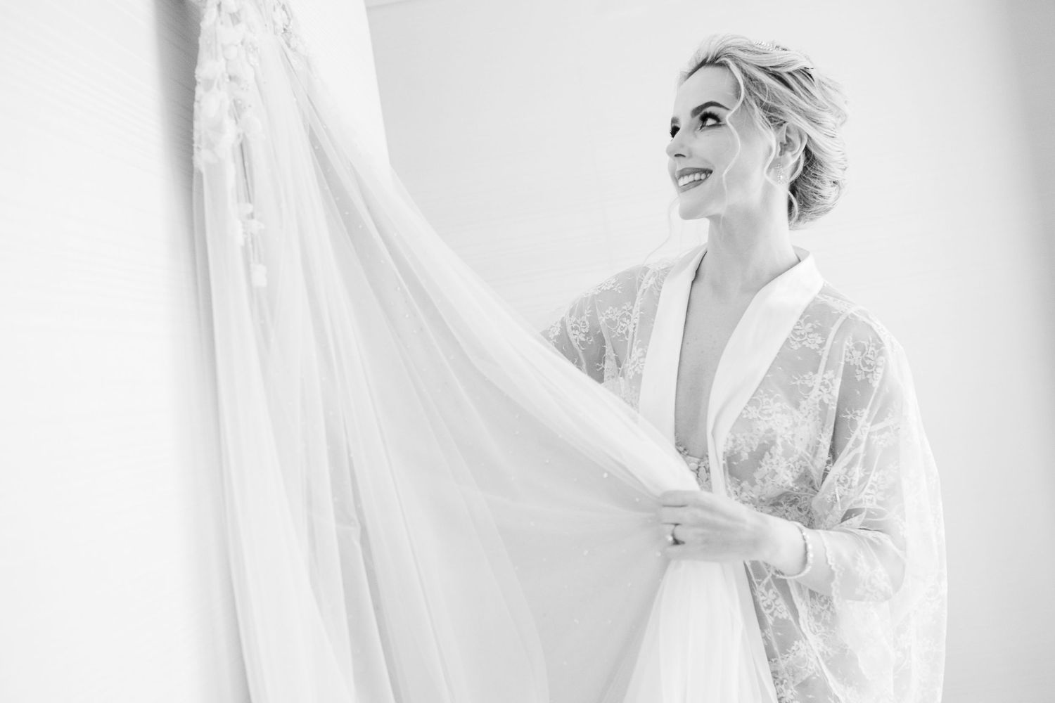 A person in a lace robe admires a flowing white wedding dress in a bright, dreamy black and white photo.