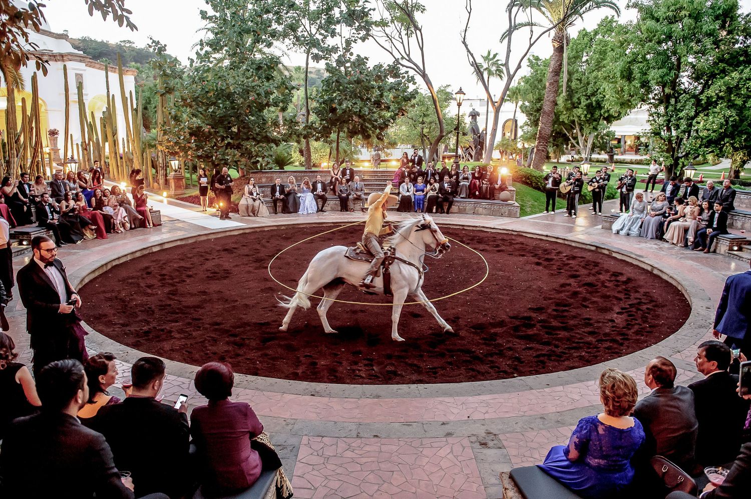 Elegant outdoor equestrian performance at a formal event, with a white horse and rider in a circular arena surrounded by guests.