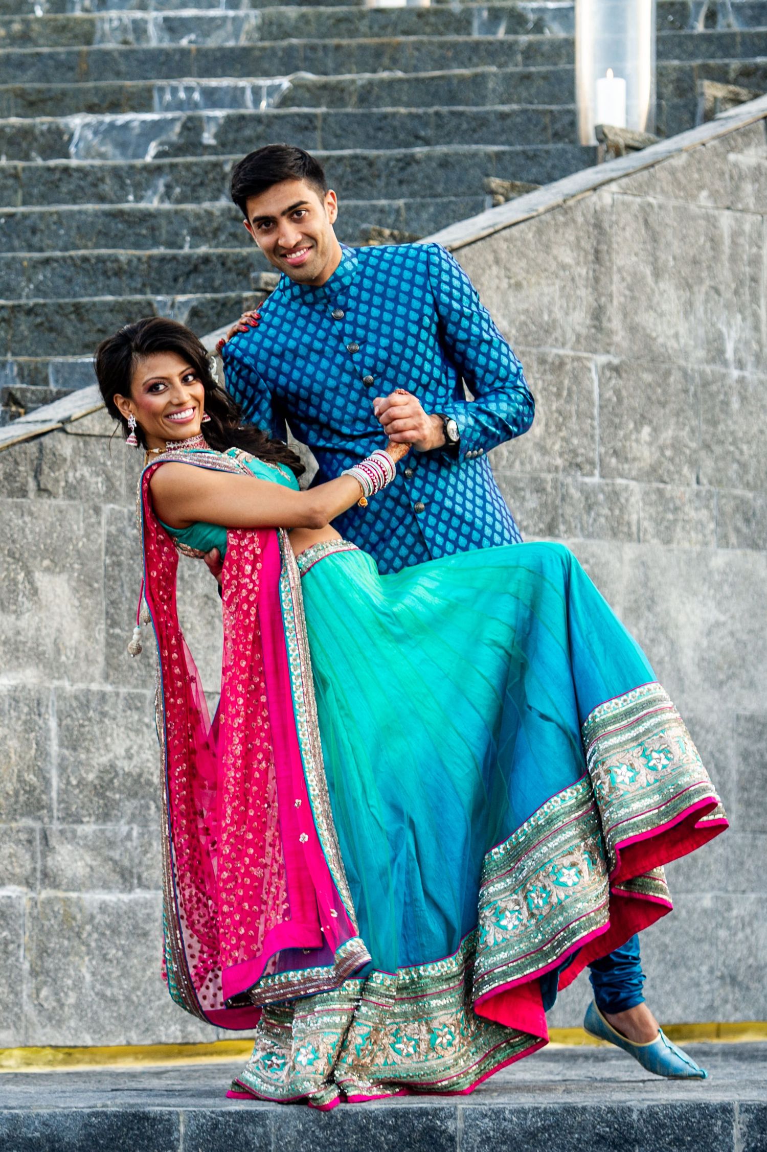 Indian couple in traditional attire, woman in pink lehenga and man in blue sherwani, posing on stone steps.