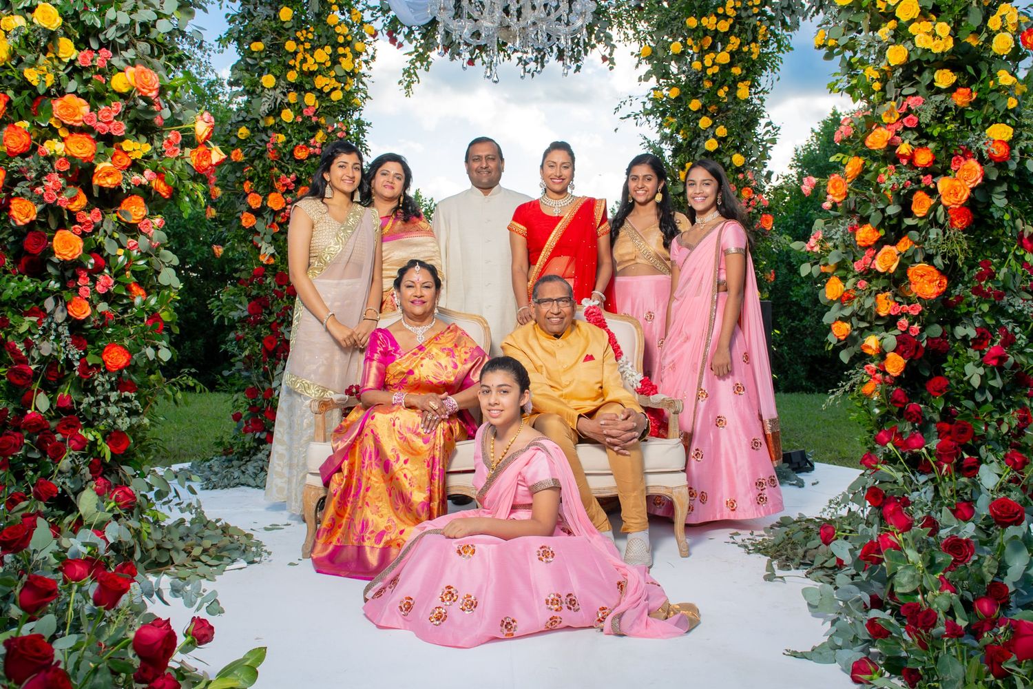 Indian family in traditional attire posing under floral arch at wedding ceremony.