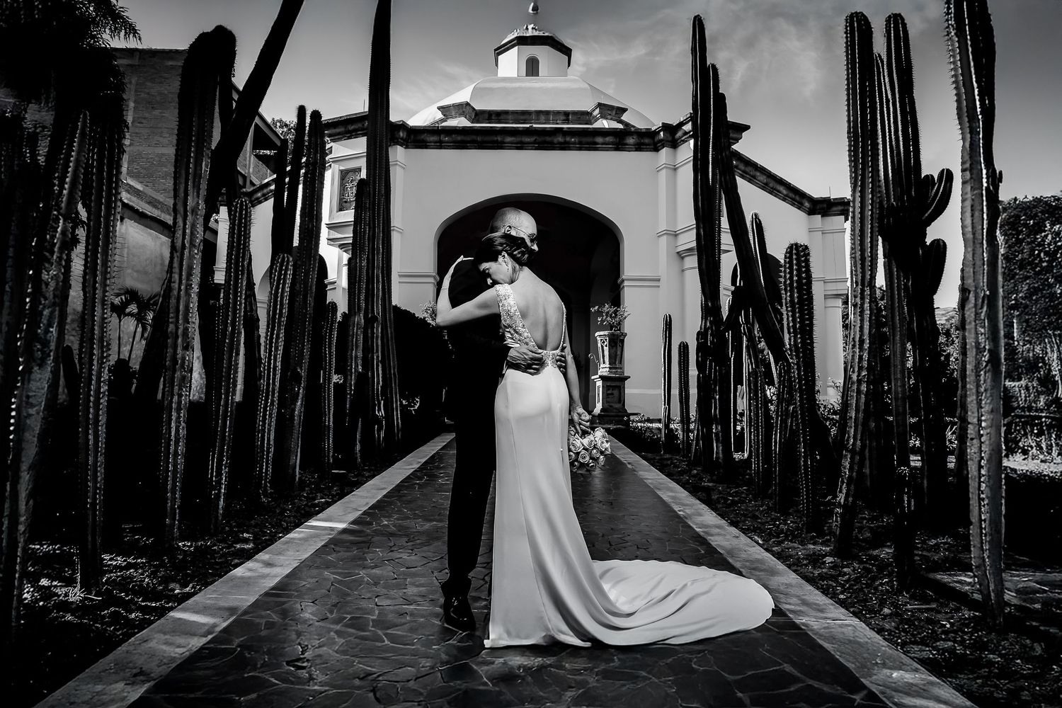 Black and white wedding photo of couple embracing among tall cacti with elegant chapel entrance backdrop.