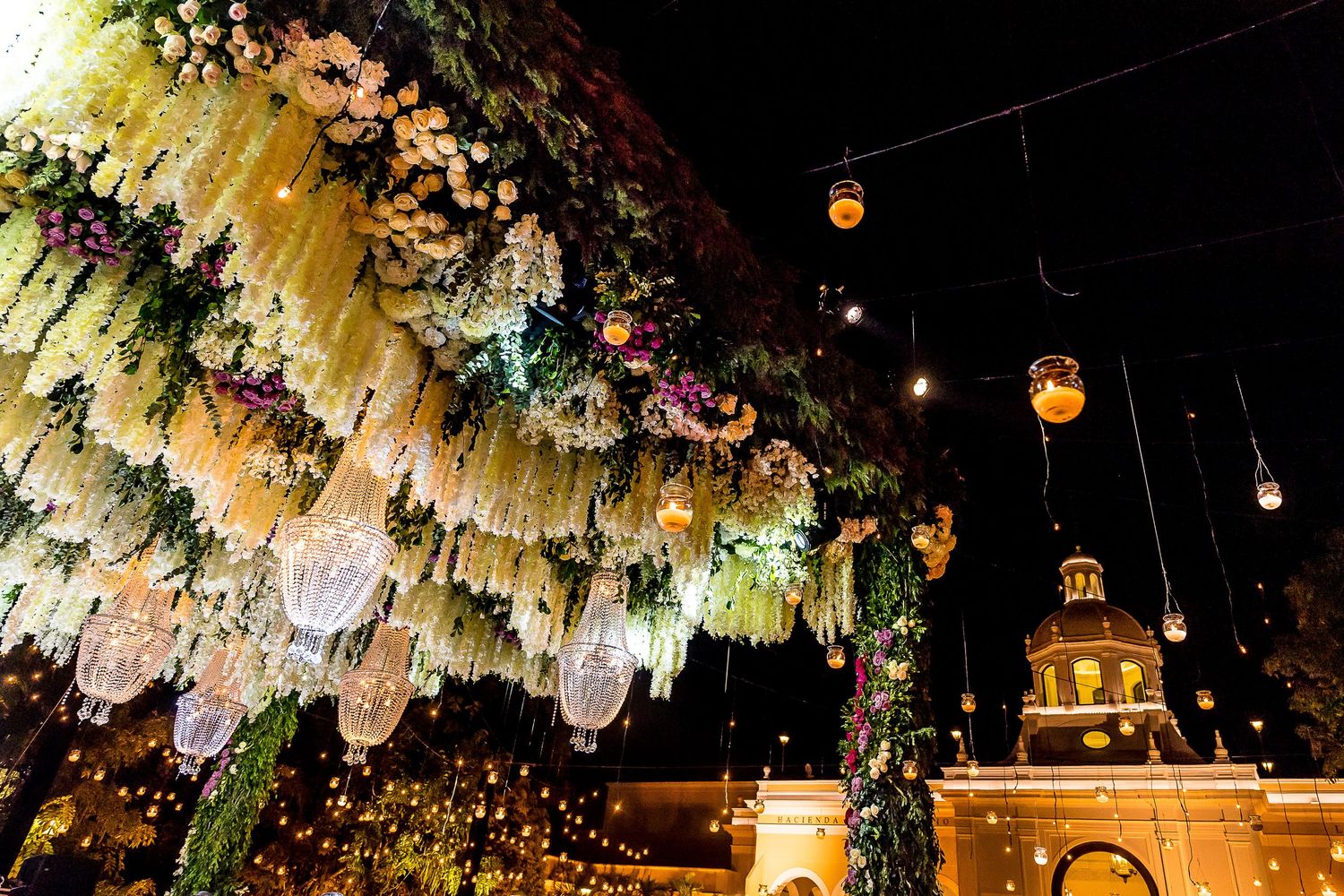 Stunning floral ceiling installation with hanging crystals, string lights, and colorful blooms at an elegant outdoor night event.