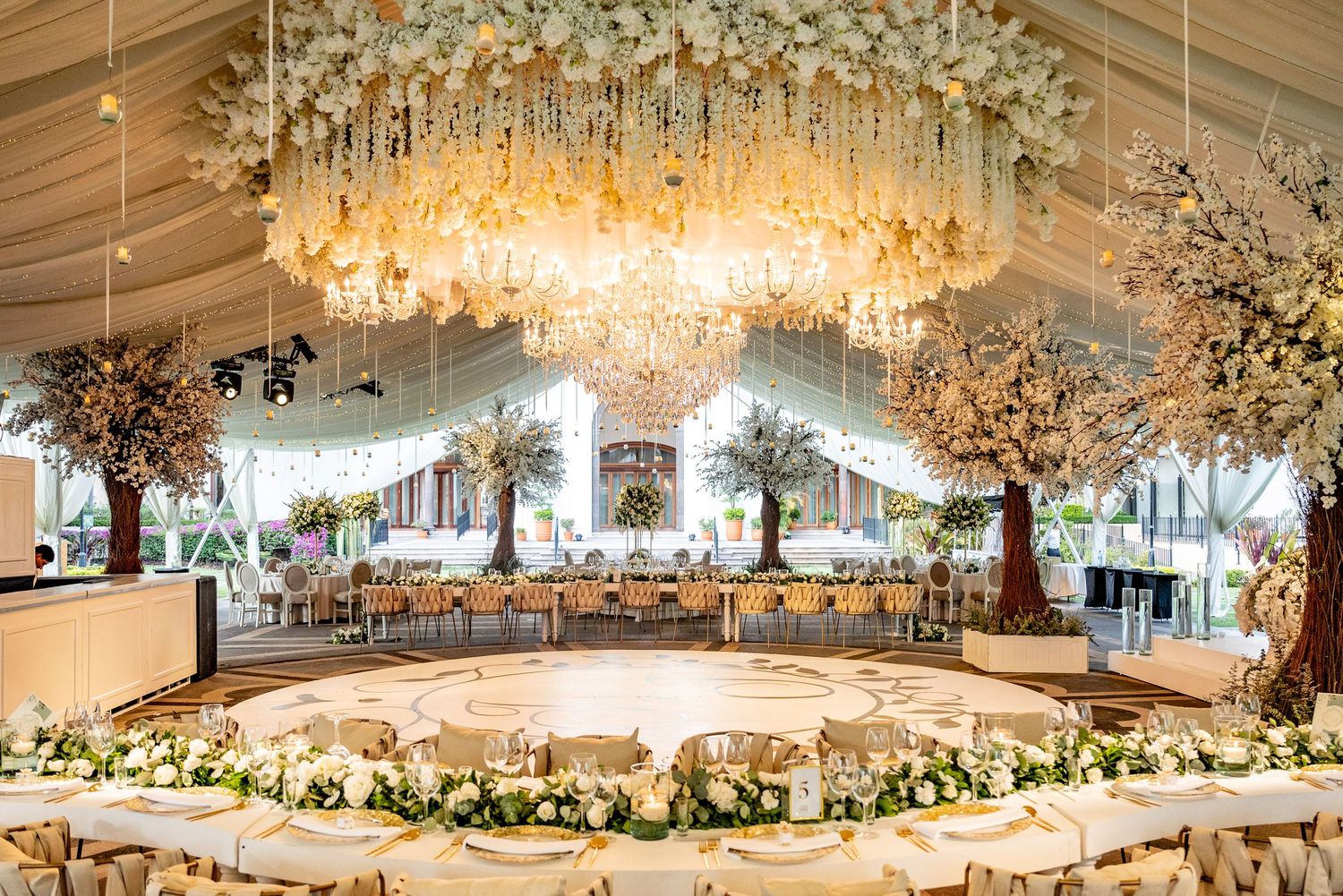 Elegant wedding reception venue with floral chandelier, crystal lights, white flower trees, and beautifully arranged dining tables.